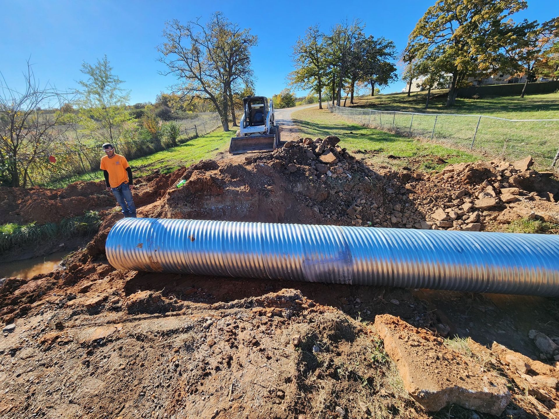Worker and excavator installing a large corrugated metal pipe in a dirt path, sunny outdoor setting.