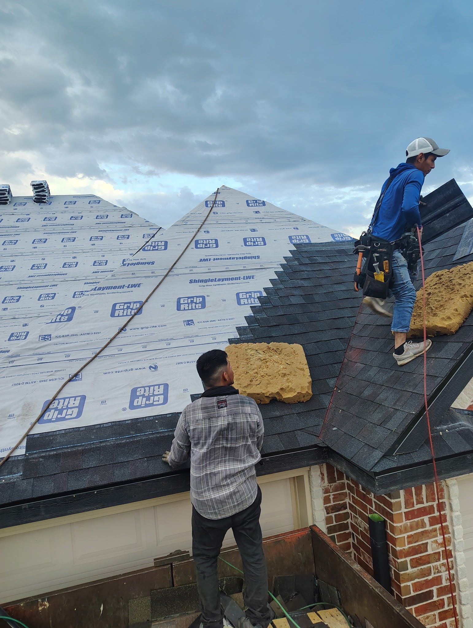 Two roofers installing shingles on a house roof; cloudy sky overhead.