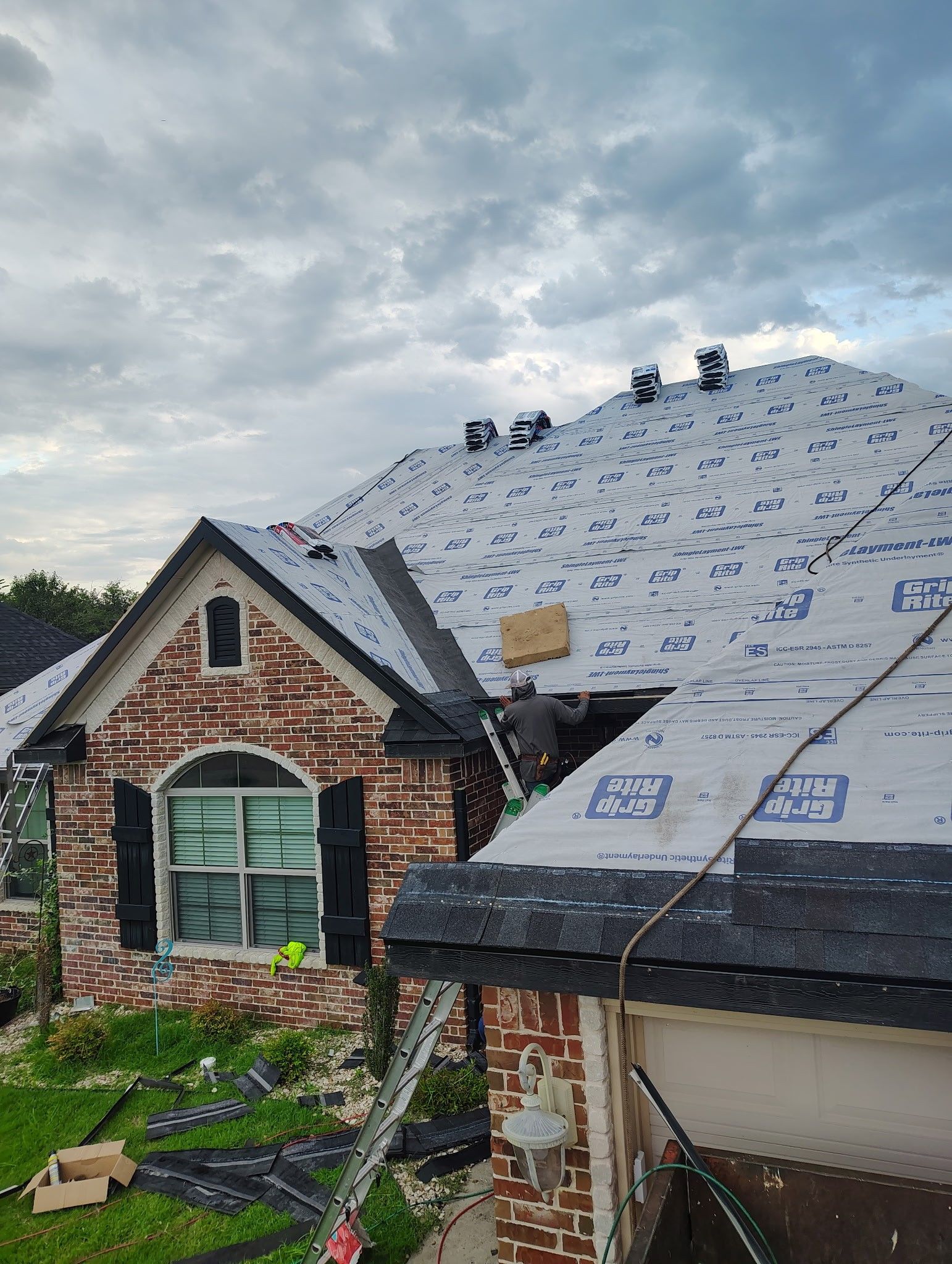 House with new roof partially installed; roofers on the roof; brick facade; cloudy sky.