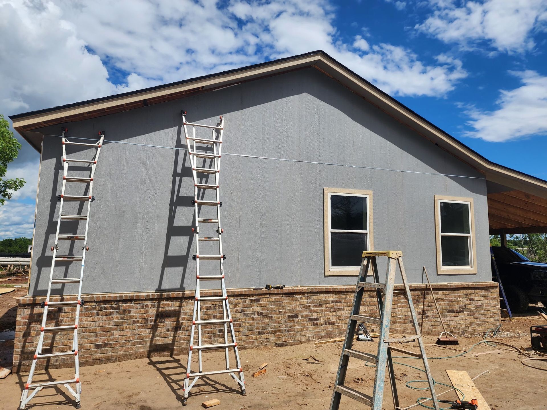 A house under construction with grey siding, brick base, windows, and multiple ladders.