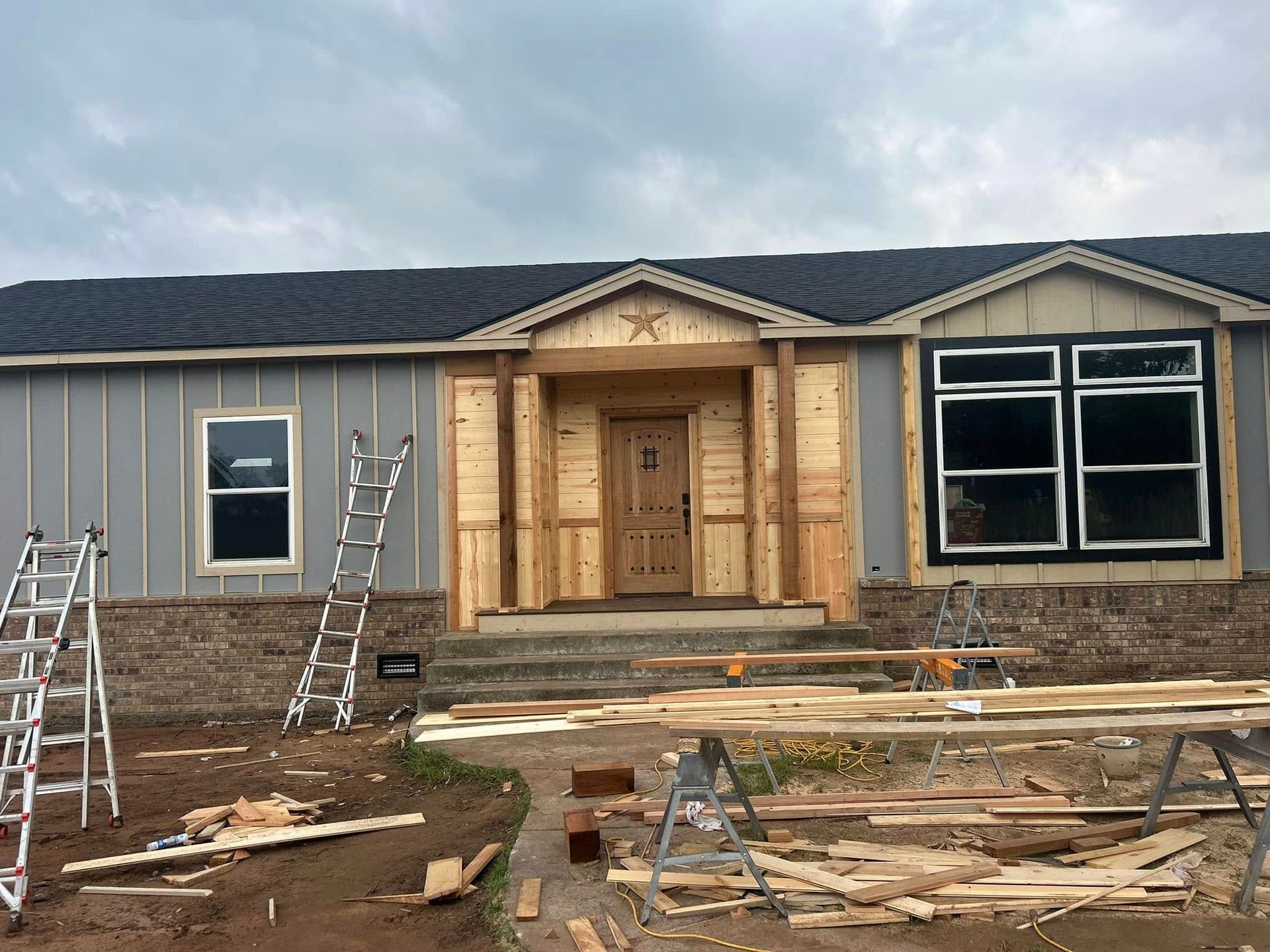 House under construction; gray siding, wooden porch, ladders, tools, cloudy sky.