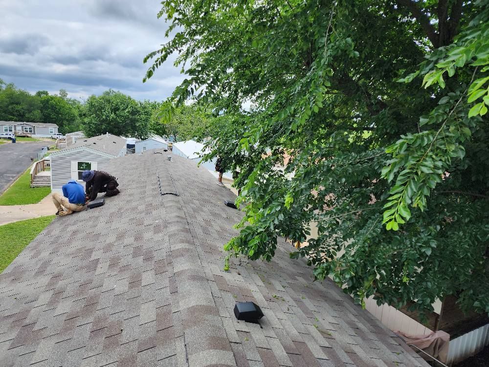 Roofers repairing shingles on a gray-shingled roof, with a green tree branch overhead under a cloudy sky.