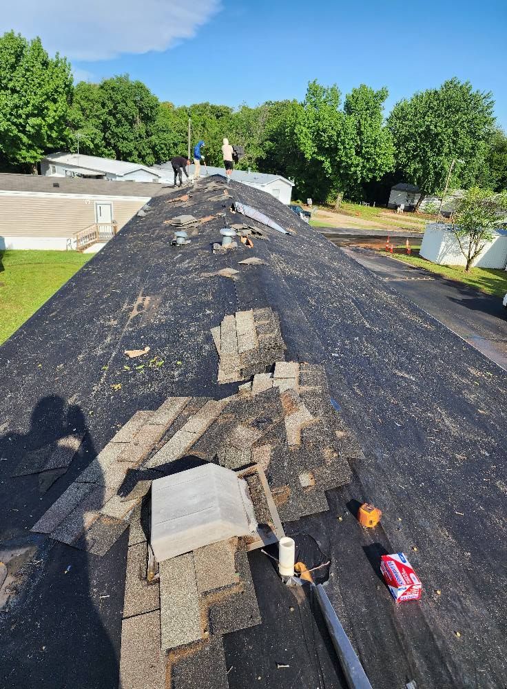 Workers on a dark roof removing old shingles. Green trees and blue sky in the background.