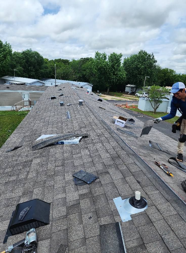 Roofer installing shingles on a gray roof. Person in blue shirt, white hat. Outdoors, overcast day.