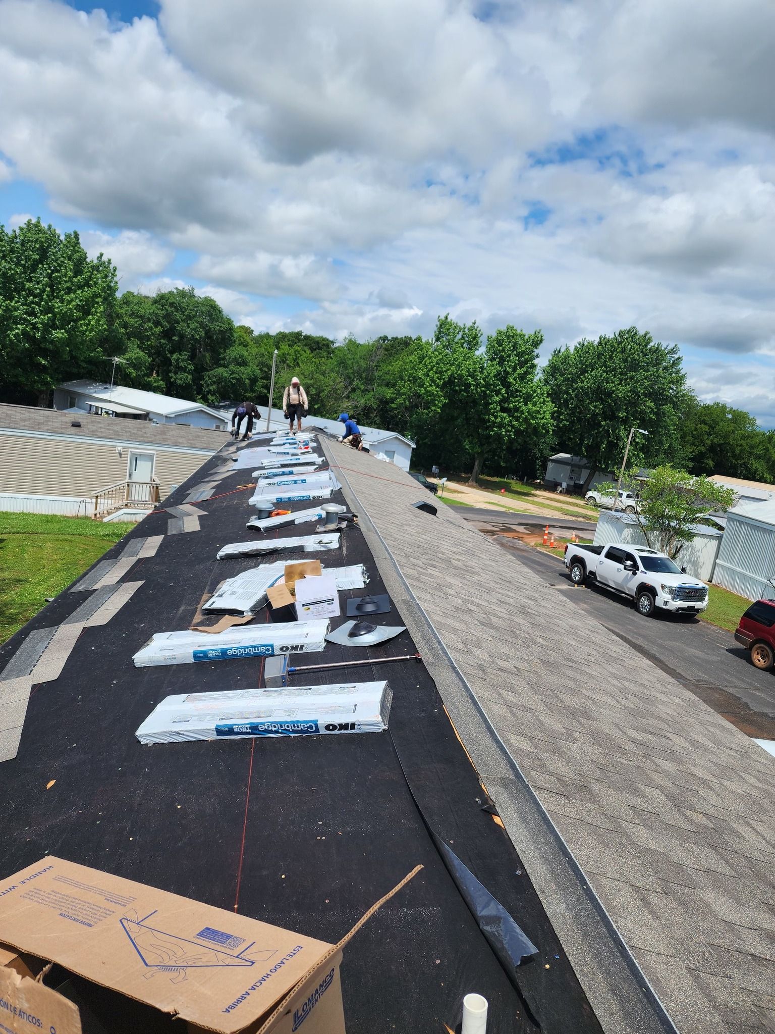 Roofing crew working on a dark shingled roof, surrounded by trees, with a few vehicles parked nearby.
