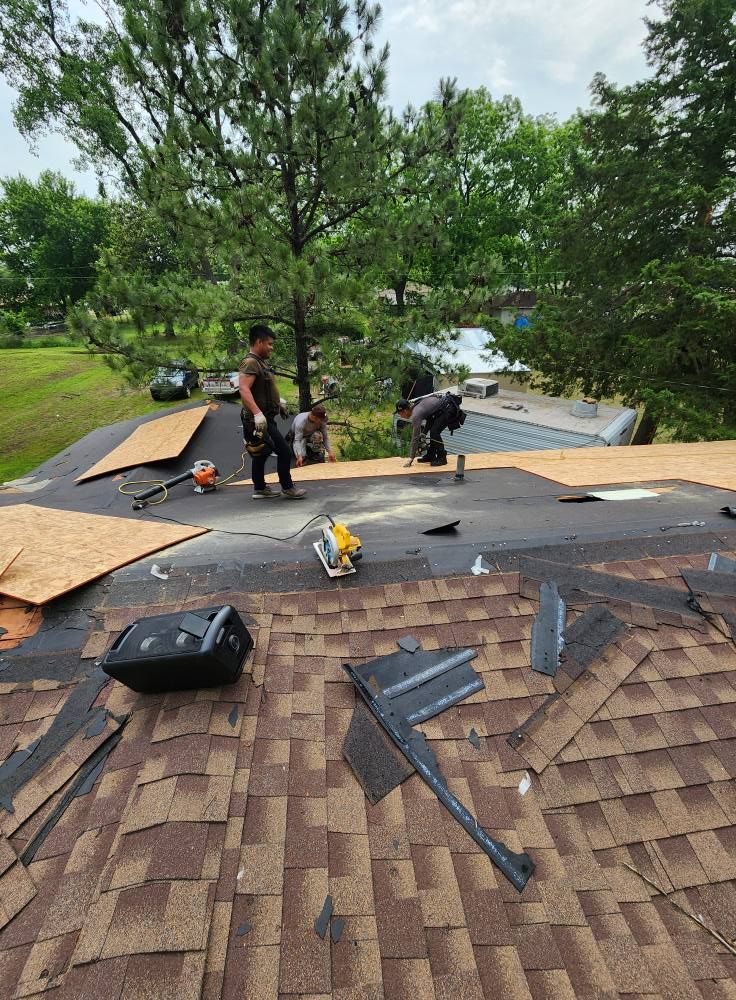 Roofers replace shingles on a house, using power tools, with trees and a body of water in the background.