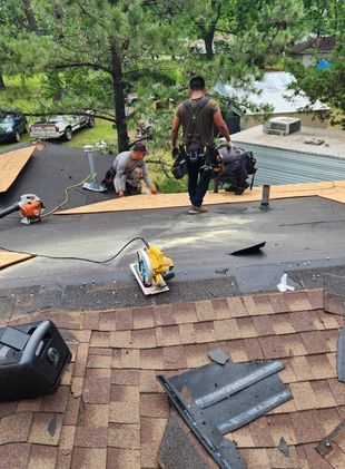 Roofers working on a shingled roof, one carrying equipment, the other installing shingles. Outdoors, trees in background.