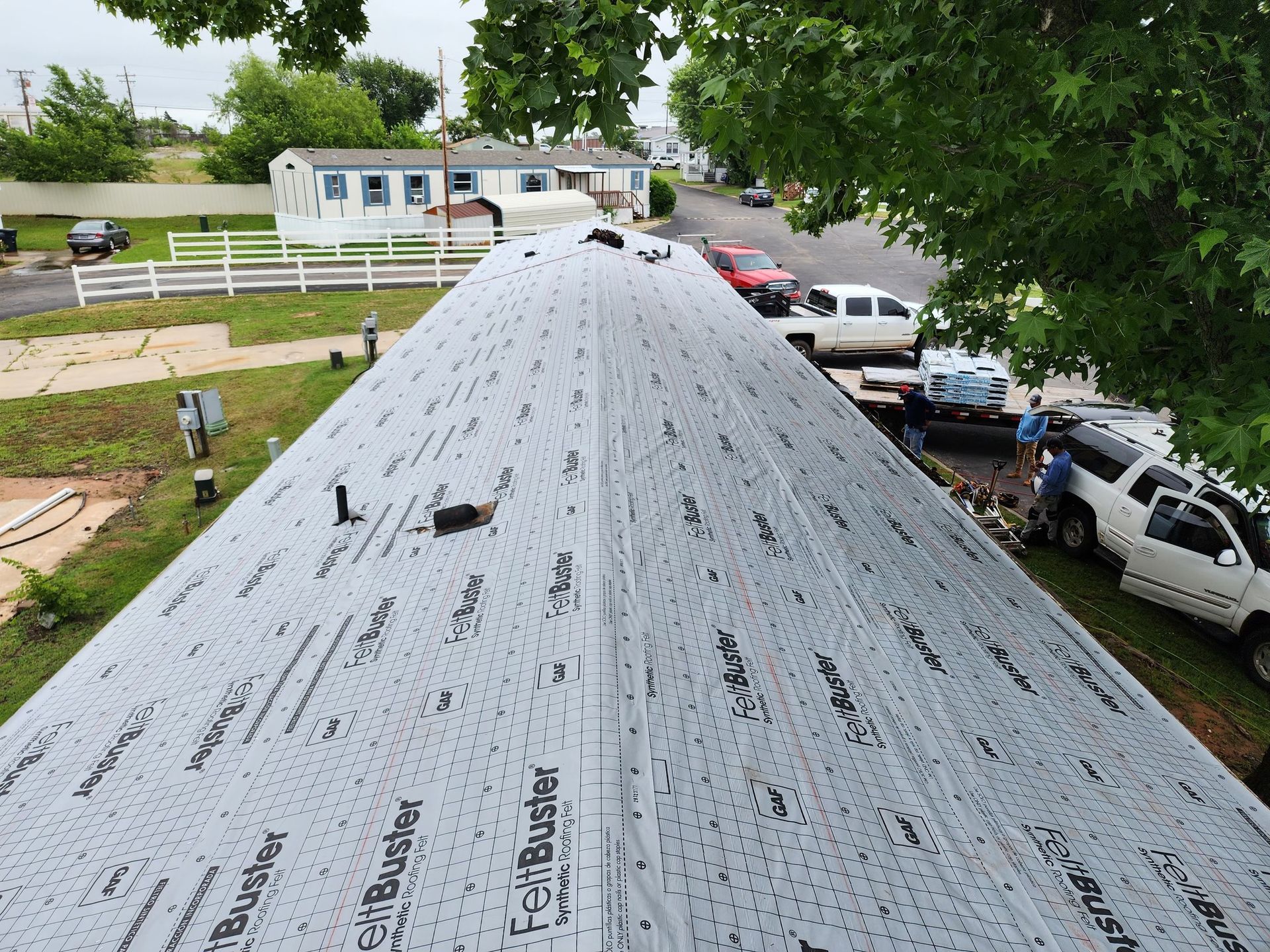 Mobile home roof with newly applied black roofing material, parked trucks in the background.