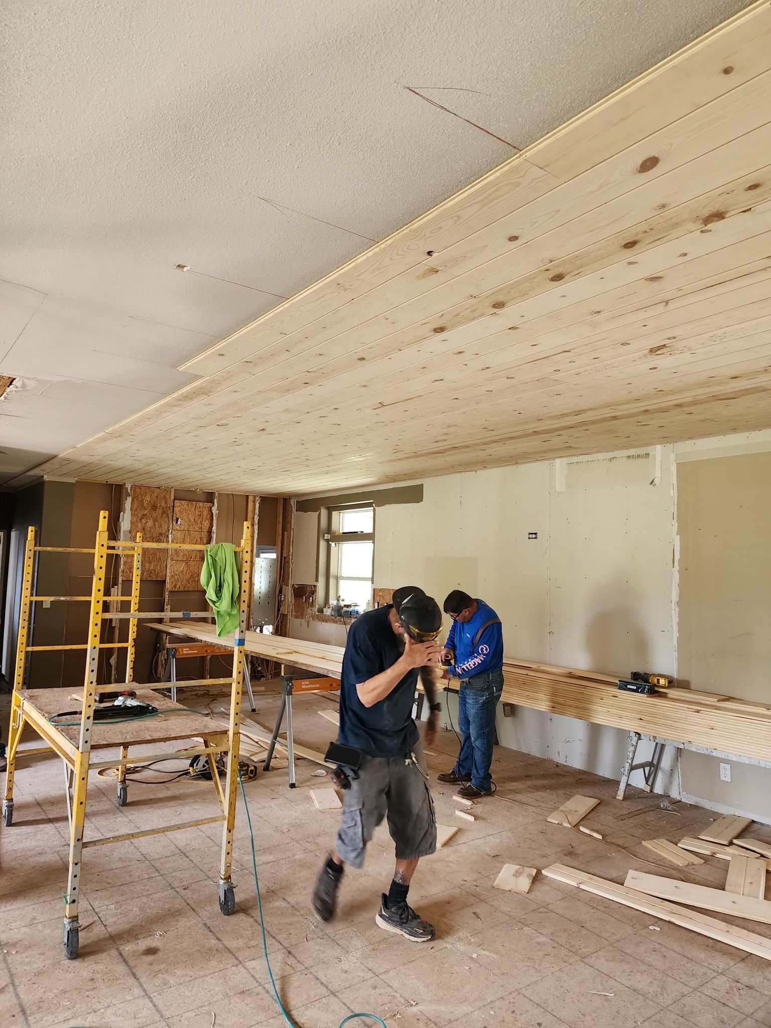 Two men installing wooden boards on a ceiling in a room; one man touching his face, tools and scaffolding visible.