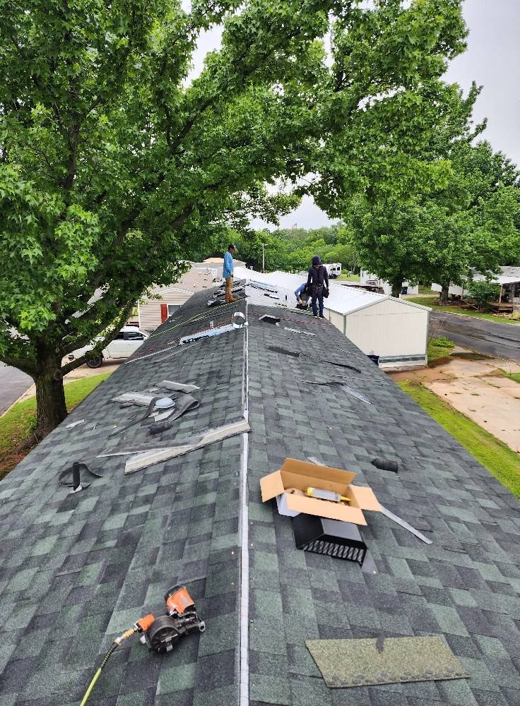 Roofers working on a house roof covered in green and black shingles, with tools and boxes present.