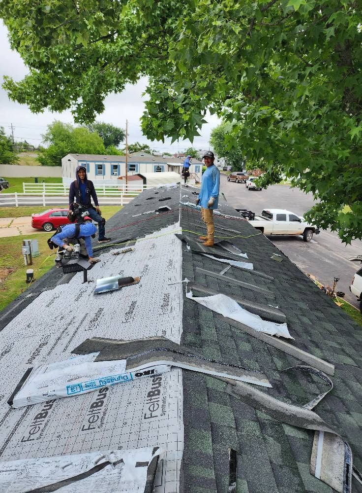 Roofers replacing shingles on a house, with some shingles removed, a street, and trees overhead.