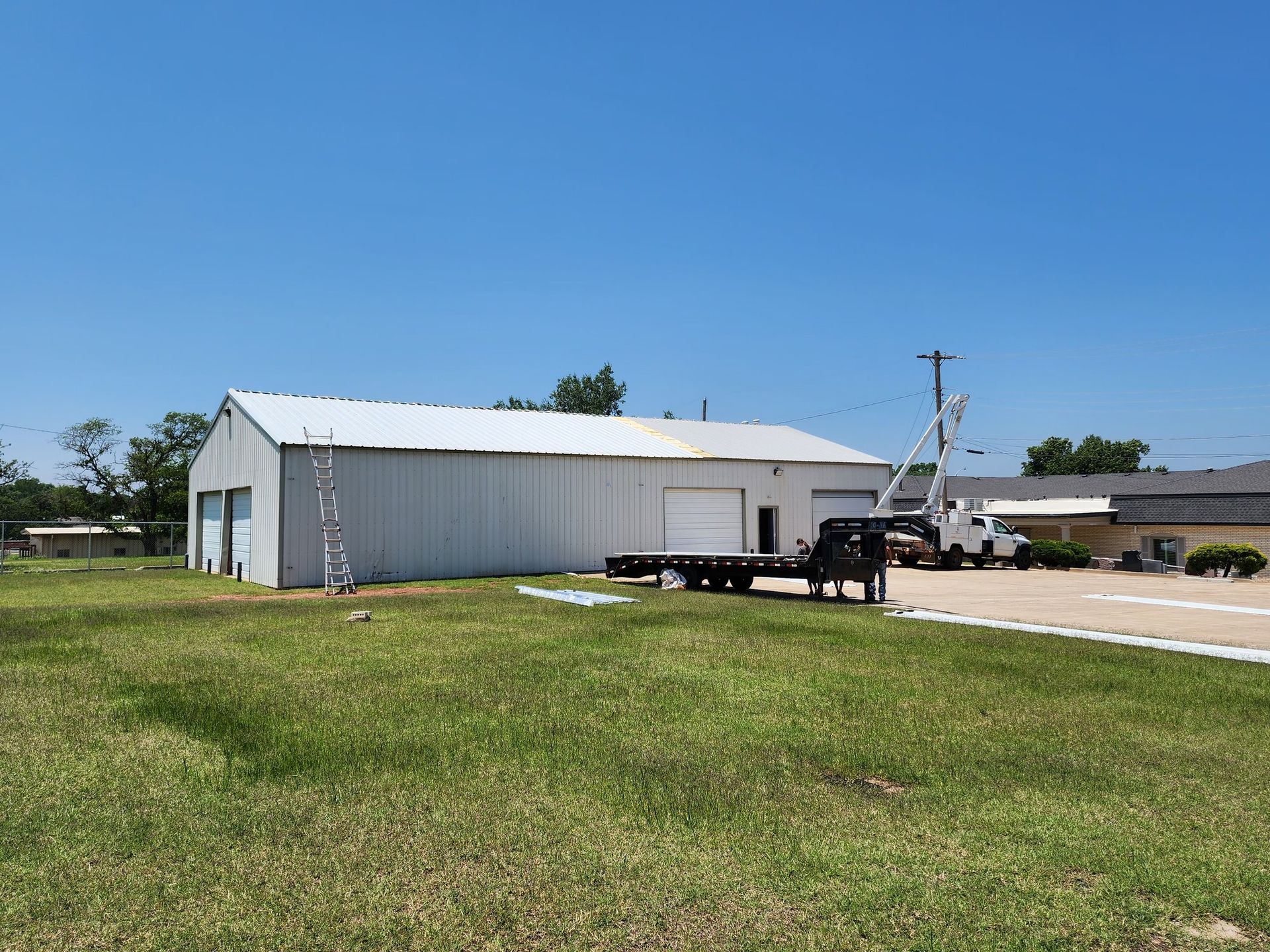 White industrial building with a flatbed trailer, utility truck, and grassy yard under a blue sky.