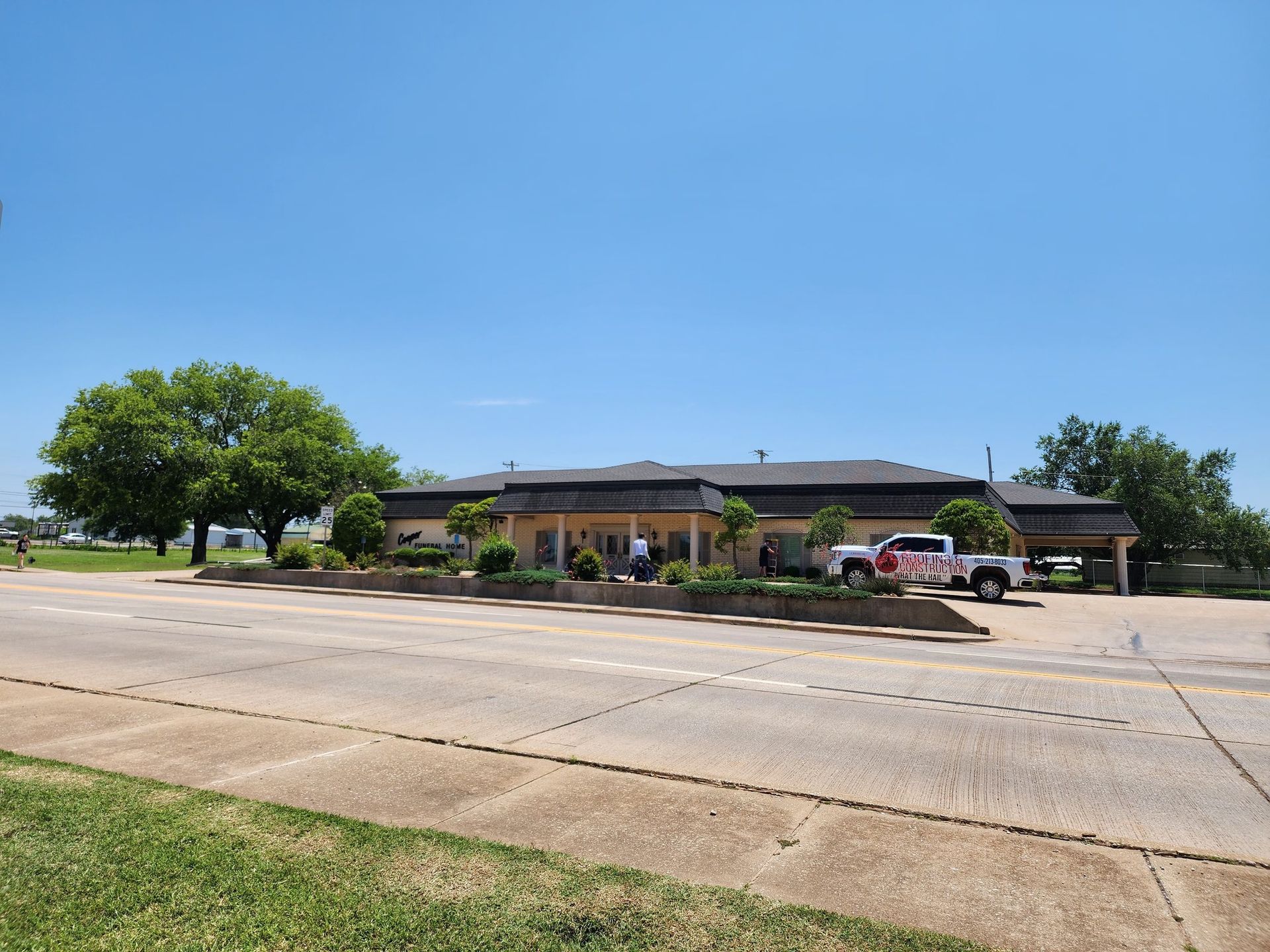 A one-story building with a brown roof, trees and a truck on the side of a road with a clear sky.