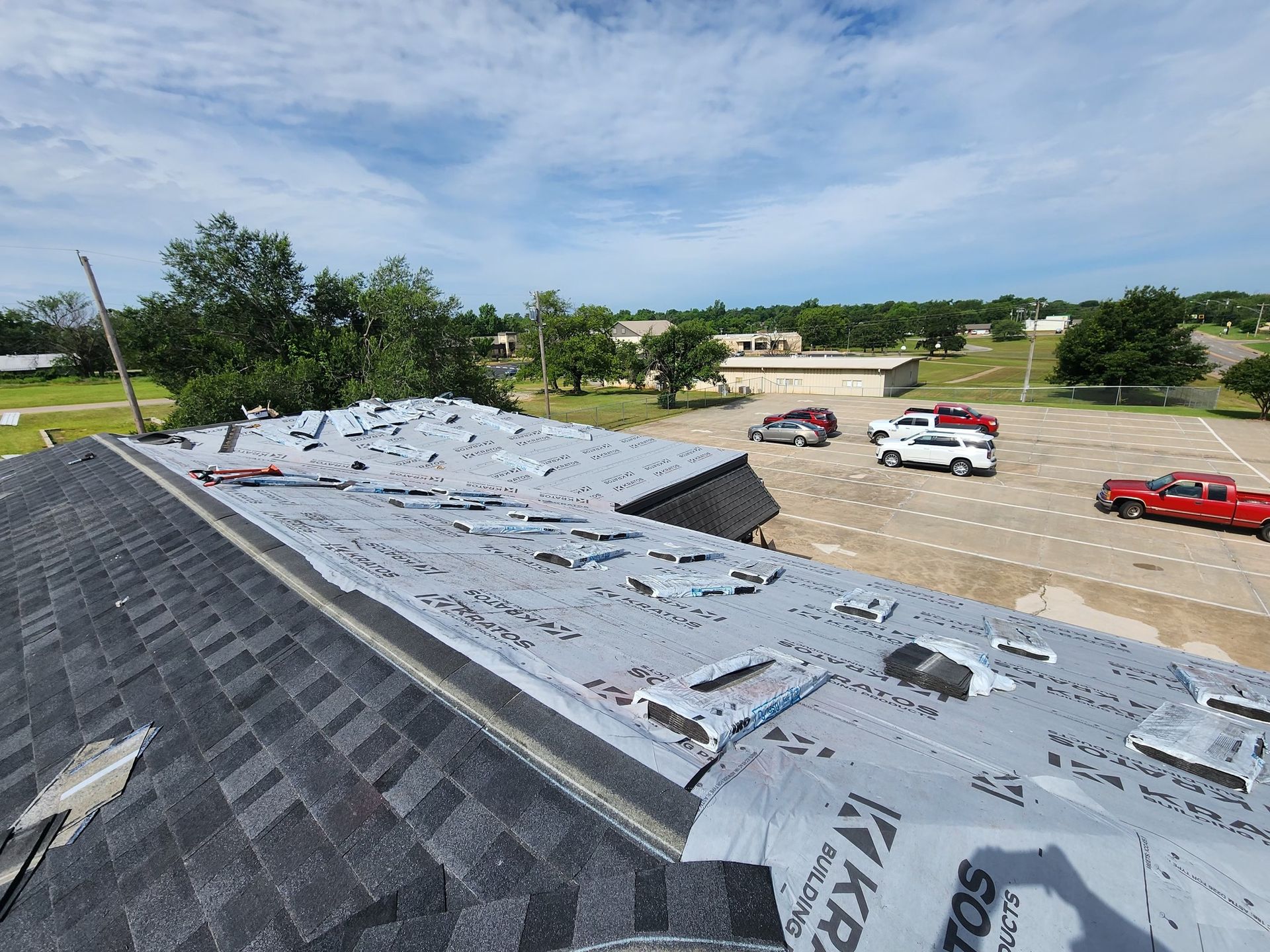 Roof being worked on, asphalt shingles partially removed, exposing underlayment. Cars parked below on a sunny day.