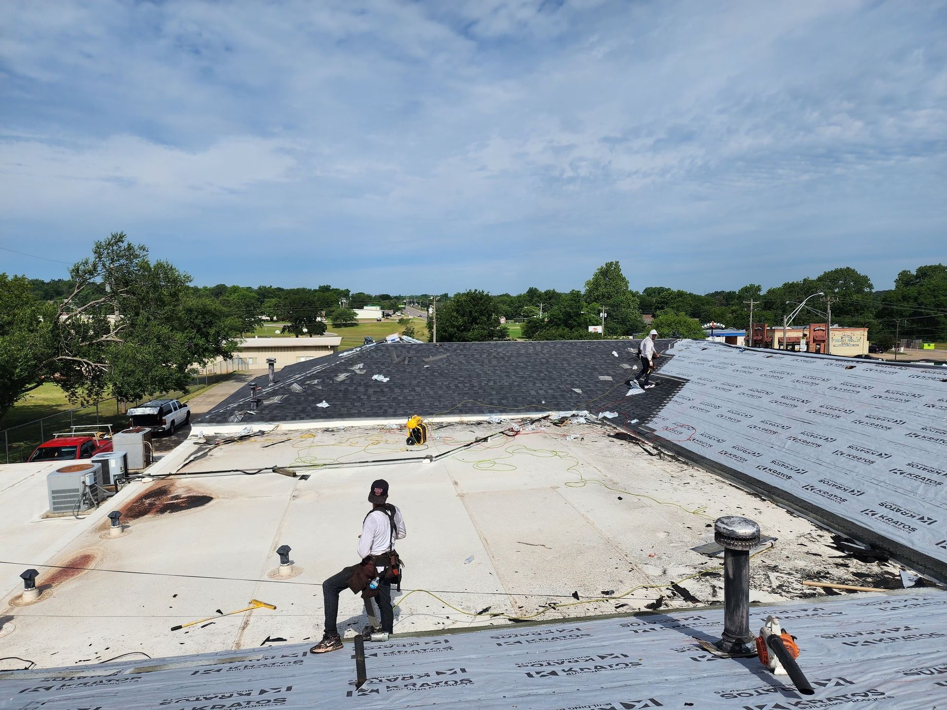 Roofers working on a building roof. One is sitting, others standing. Blue sky, trees, and other buildings in the background.