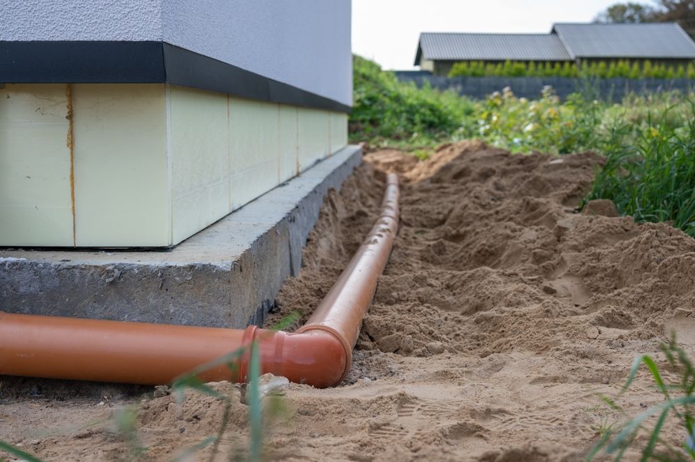 Orange drainage pipe along a house foundation beside a sandy trench in a yard