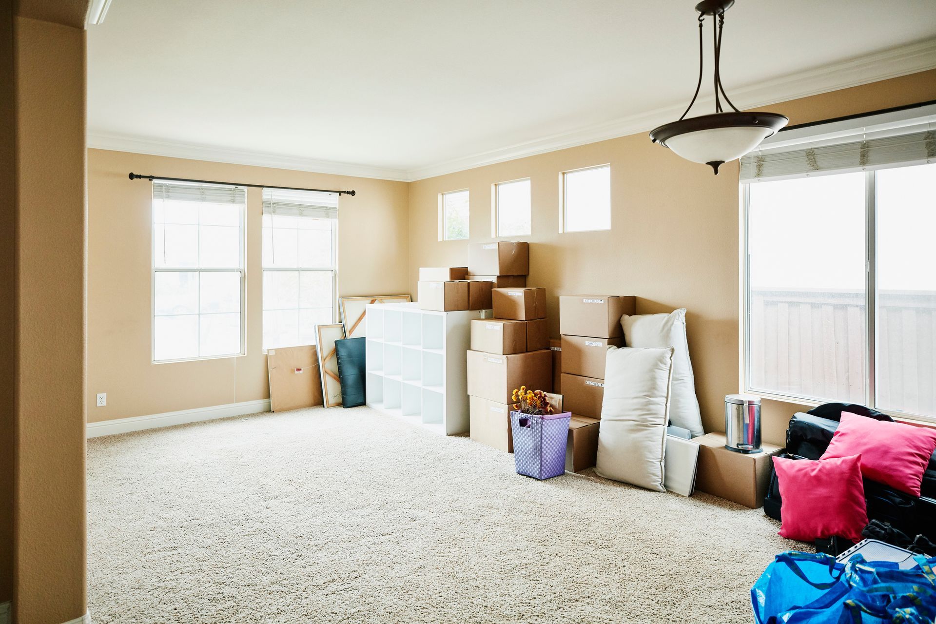 Empty room with beige walls and carpet, full of stacked moving boxes and some bags.