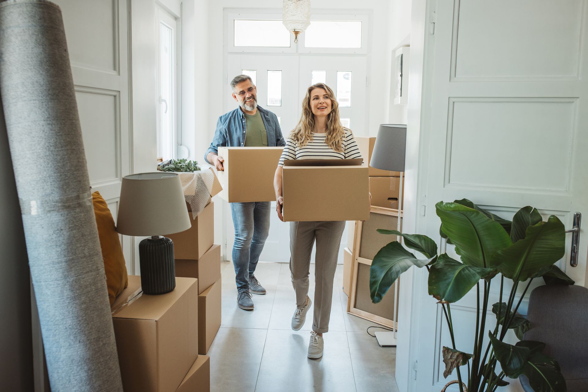 Couple carrying moving boxes into a bright hallway, smiling, with stacked boxes and plant.