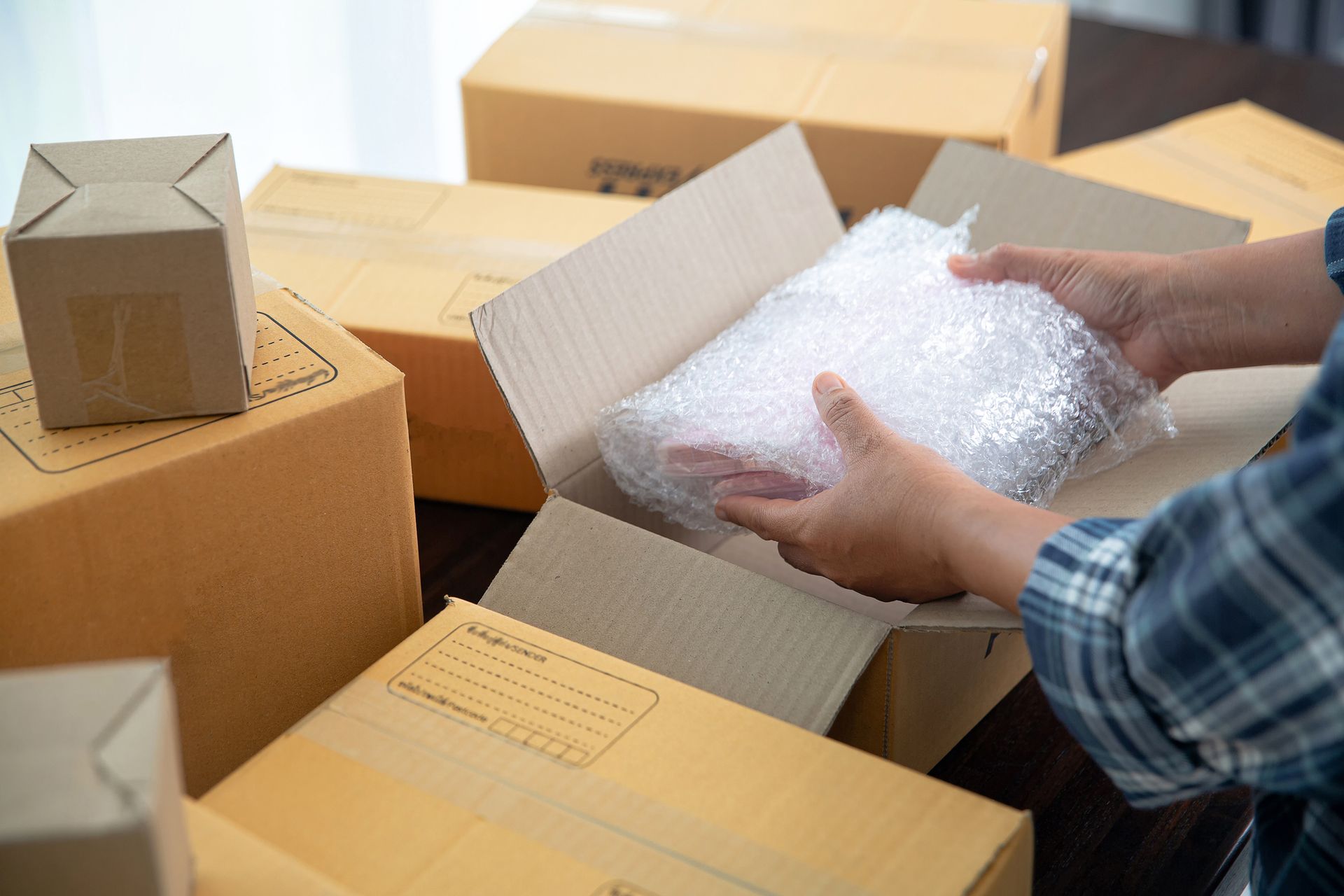 Hands packing a bubble-wrapped item into a cardboard box surrounded by other packages on a dark surface.