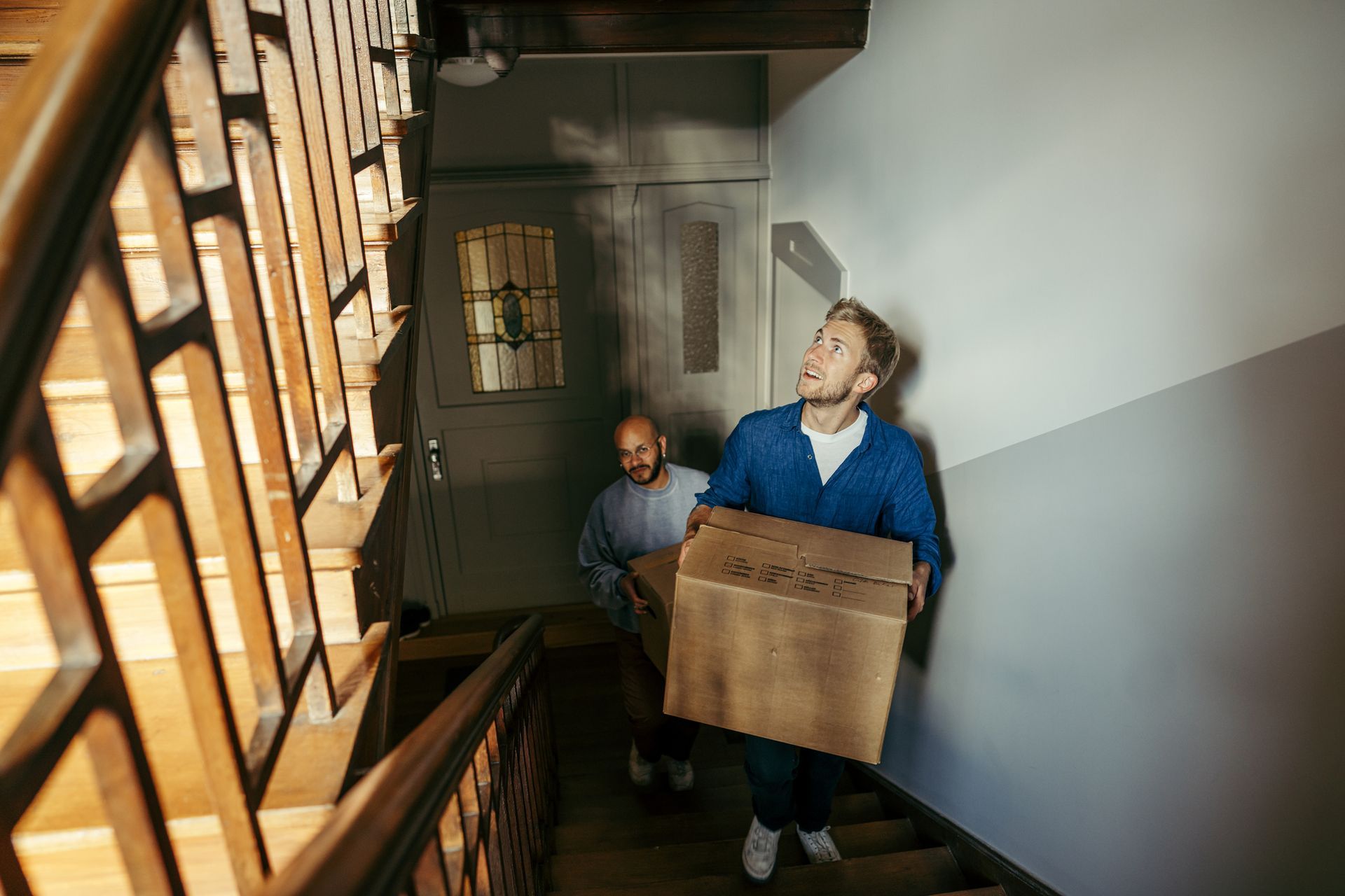 Two people carrying boxes up a wooden staircase inside a building. One looks up.