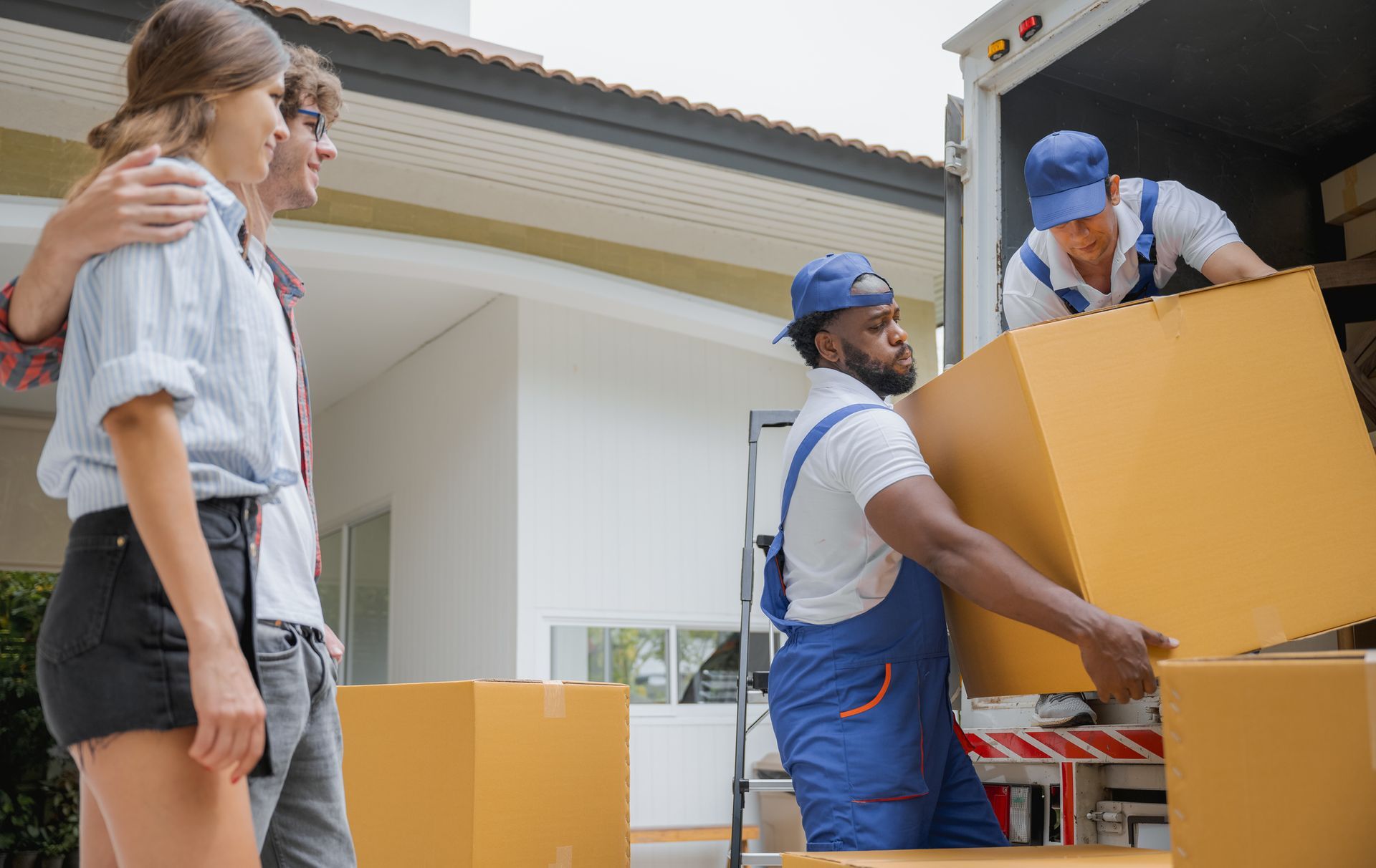 Couple watches movers unloading boxes from a truck near a house.