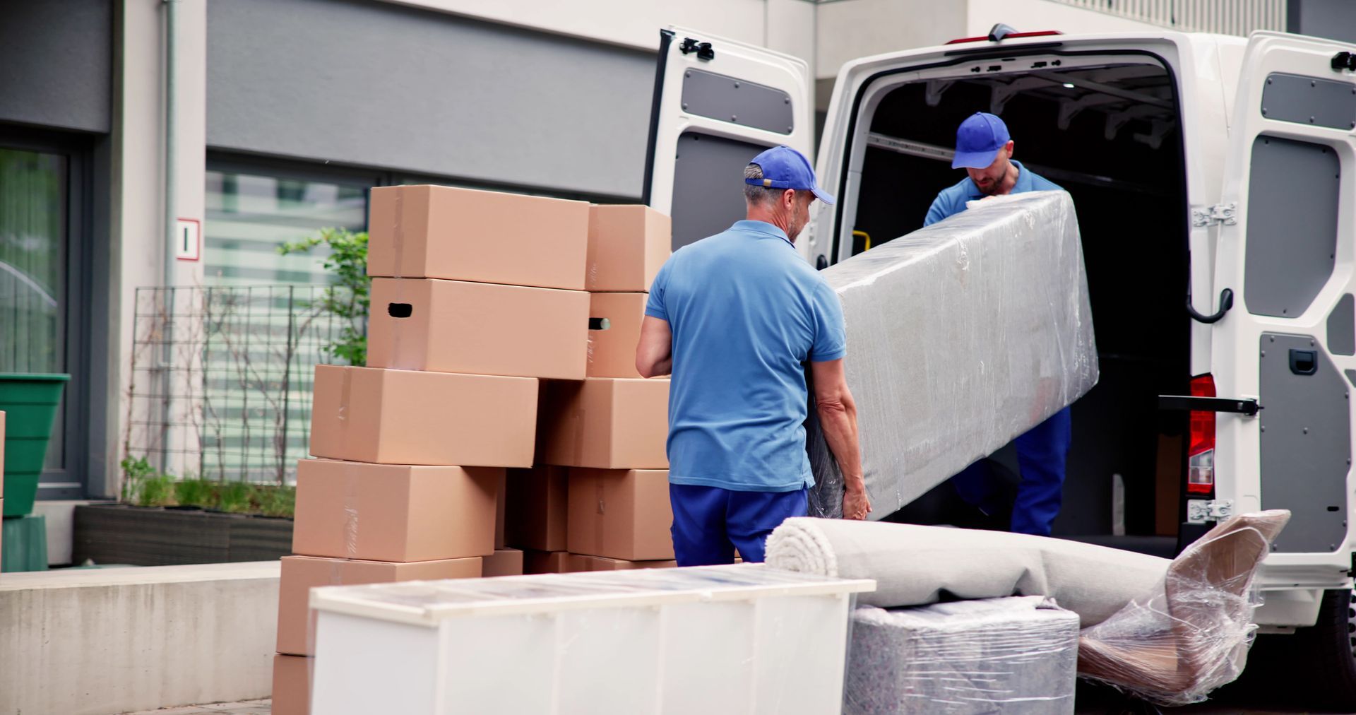 Movers loading a large upholstered furniture piece into a white van; boxes piled nearby.