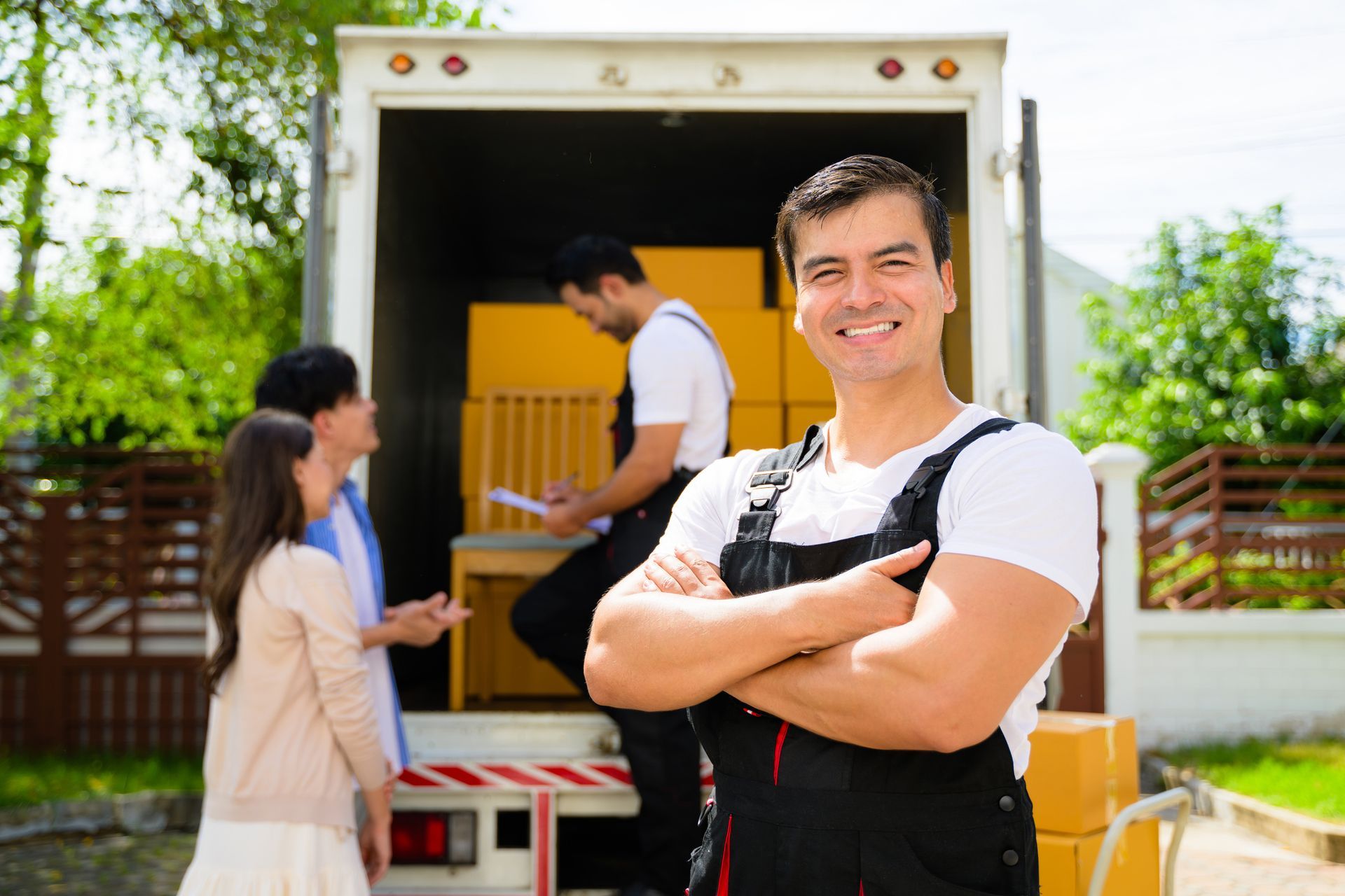 Smiling mover in overalls stands by truck; couple watches as another mover loads boxes.