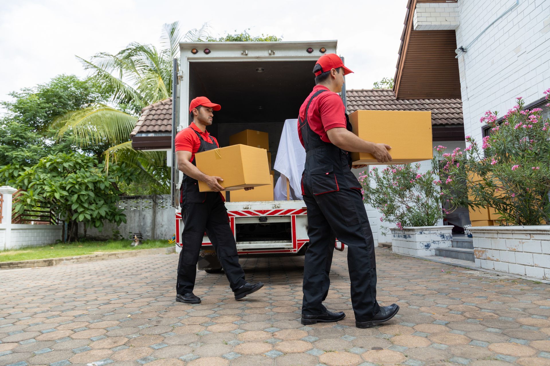 Two movers in red and black uniforms carrying boxes from a truck to a house.