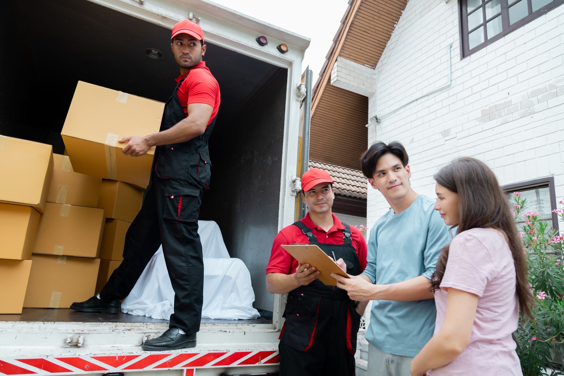 Movers loading boxes into a truck while a couple reviews paperwork outside a house.