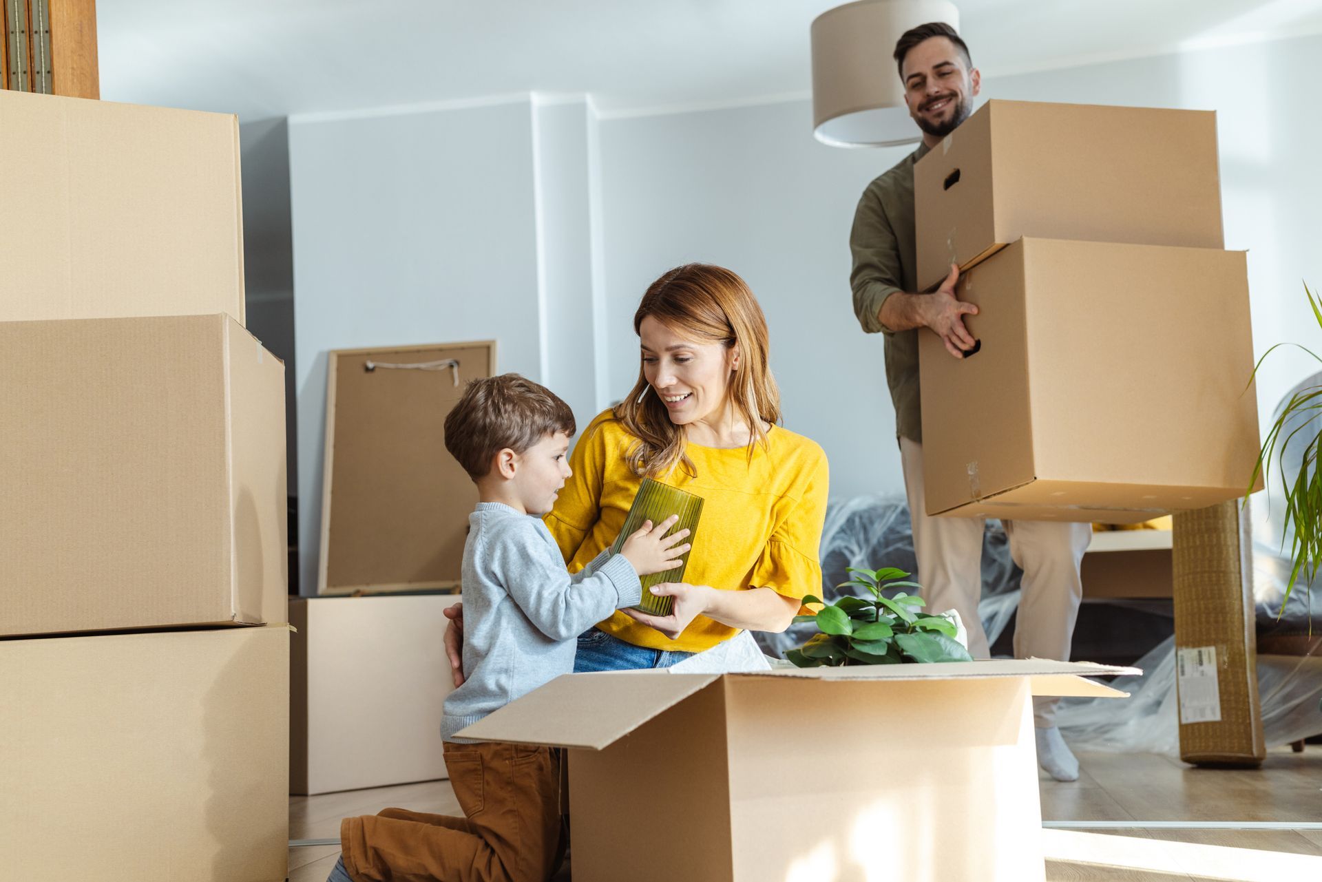 Family unpacking boxes in new home: woman smiles, boy holds plant, man carries boxes.