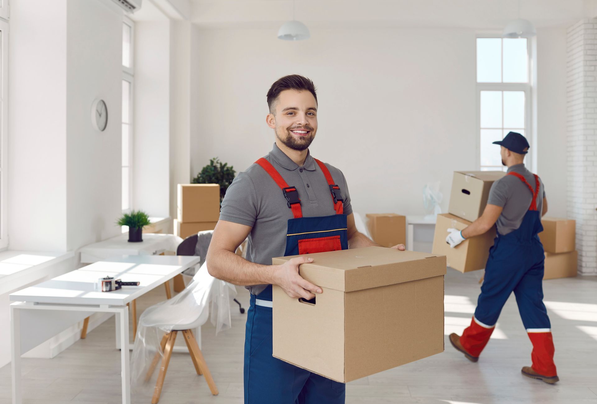 Movers carrying boxes in an empty, bright room. One smiles at the camera.