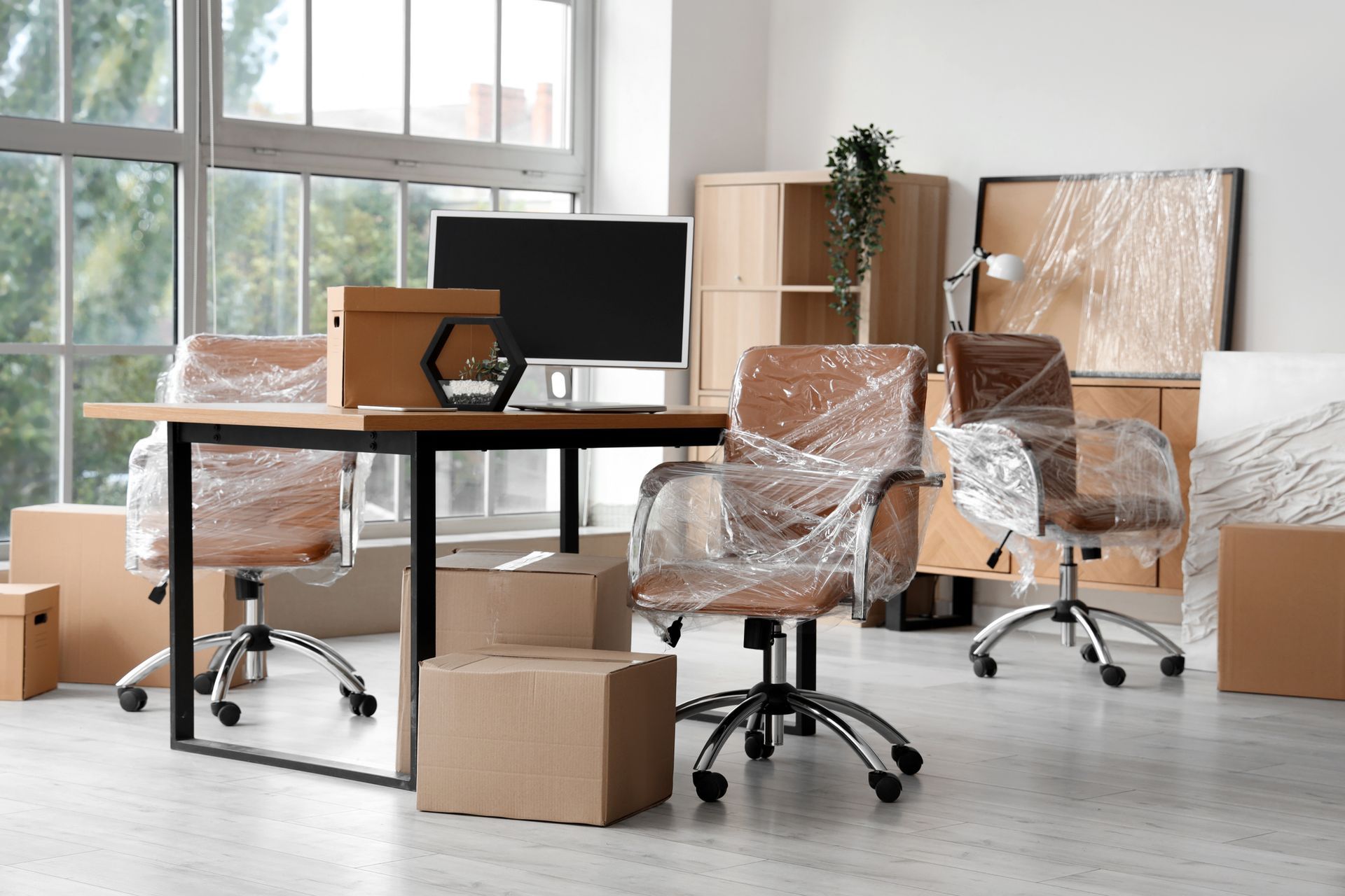 Office furniture wrapped in plastic, with boxes, ready for moving. Bright room with window.