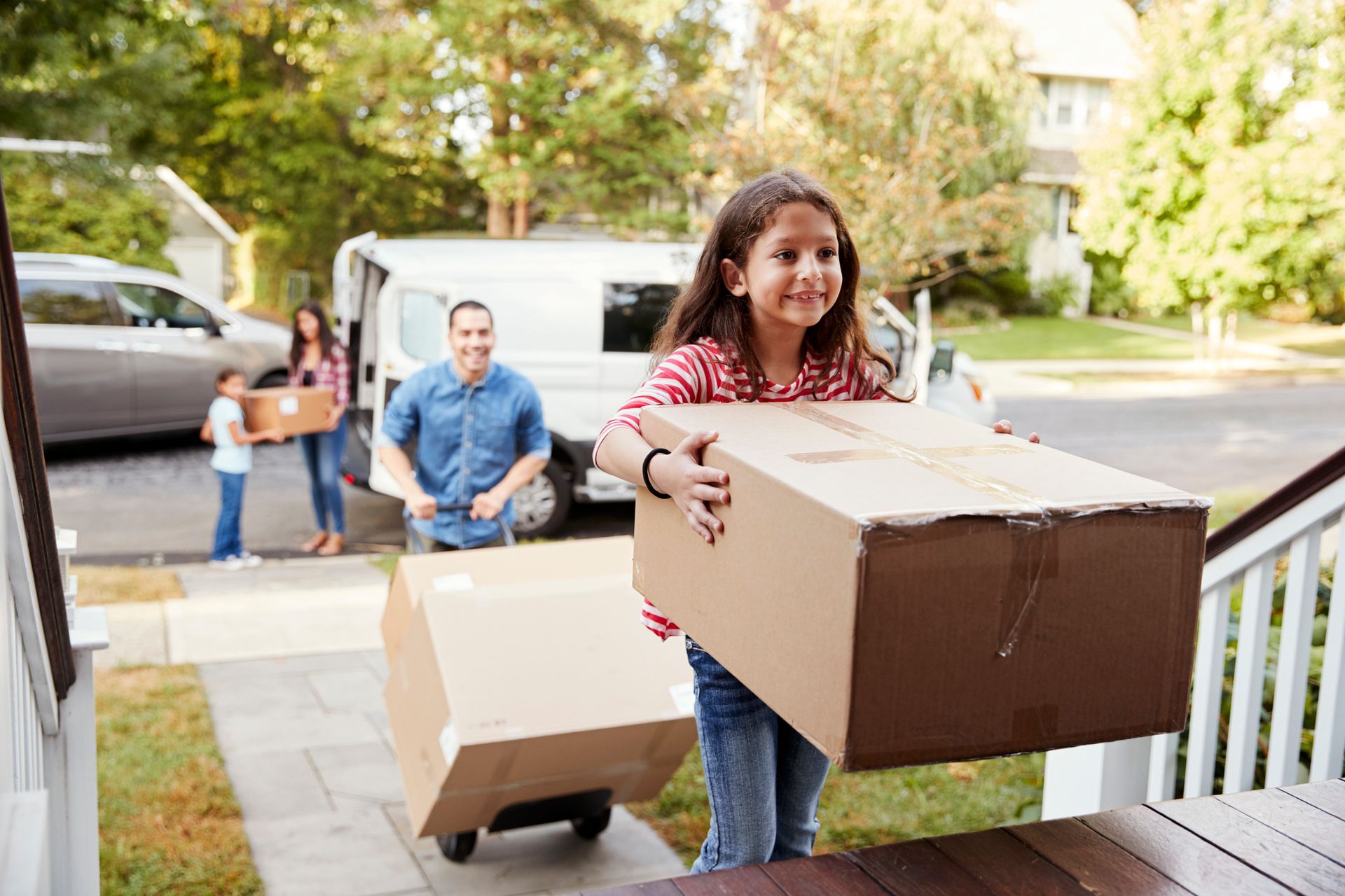 Family moving boxes from a van into a house. Girl carrying a box, others unloading. Sunny day.