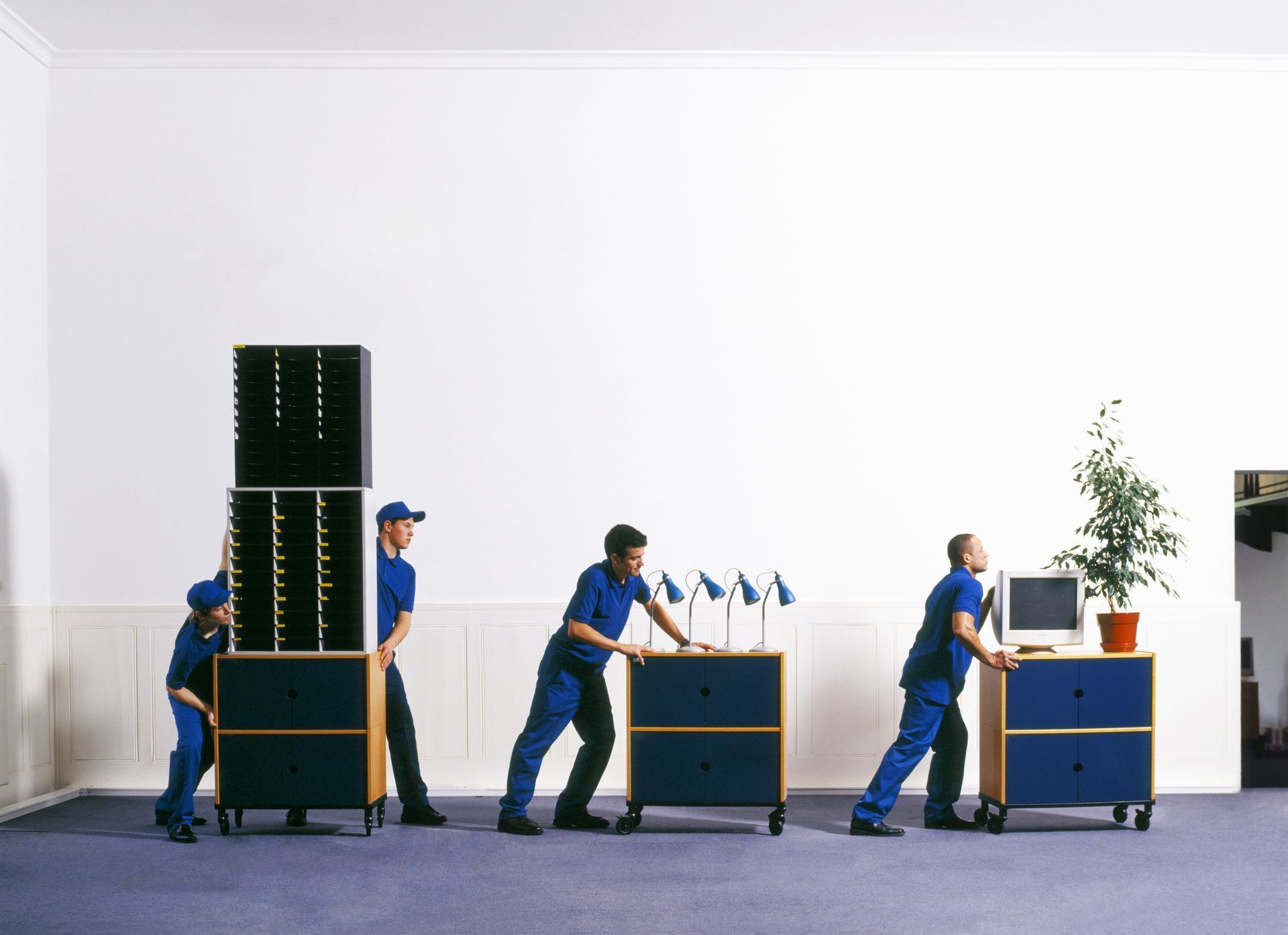 Four people in blue uniforms pushing office furniture on wheels across a blue carpet, against a white wall.