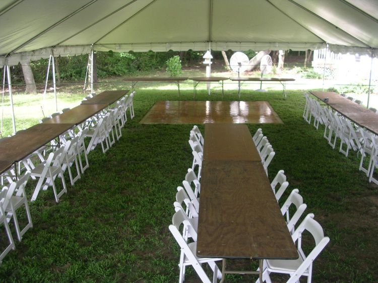 Inside a tent, long tables with white chairs arranged for an event on a grassy area.