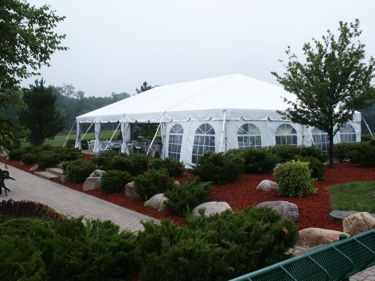 White tent set up on red mulch, surrounded by green bushes and trees, a stone pathway visible.