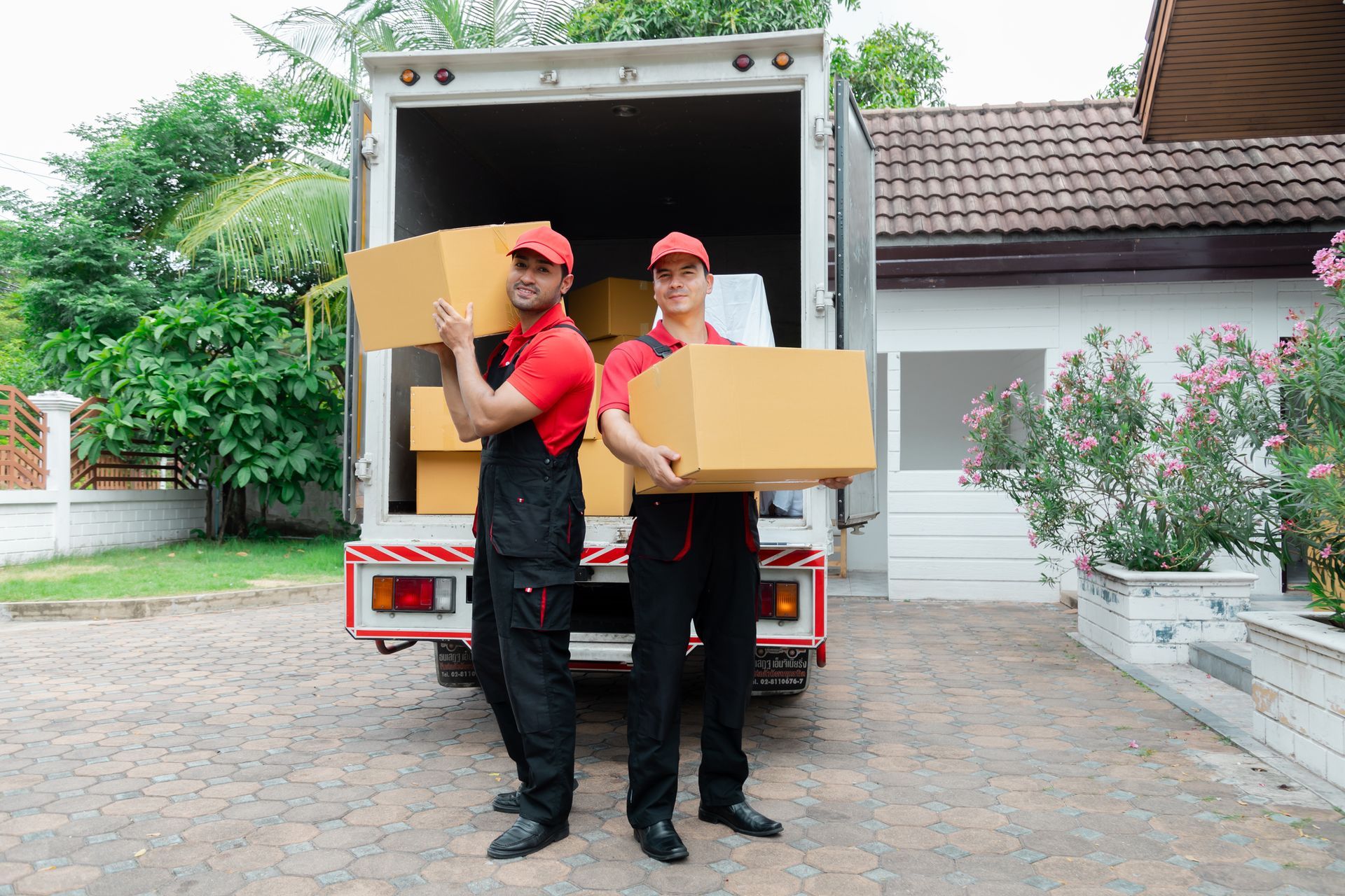 Two movers in red shirts and caps carry cardboard boxes from a truck into a house.