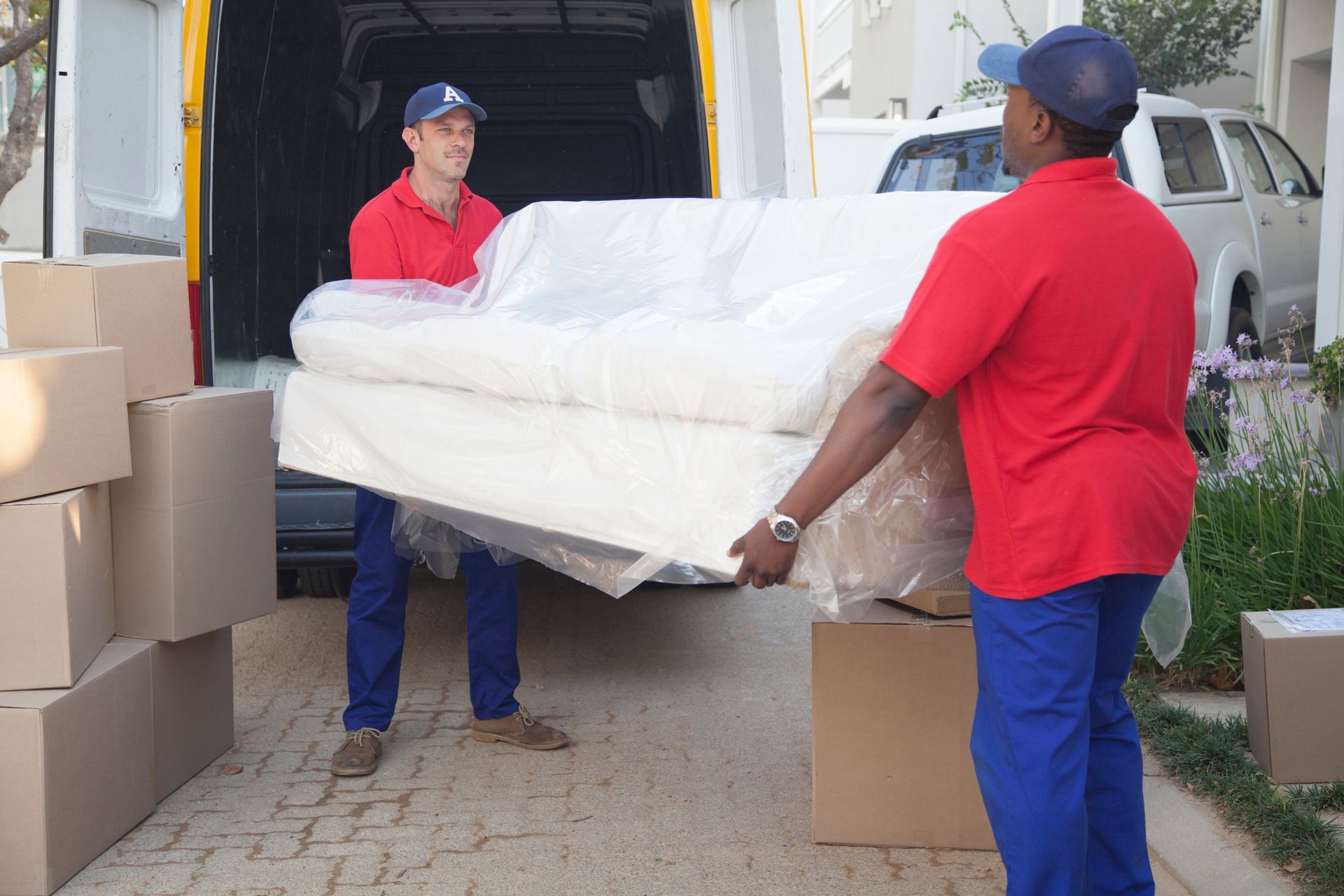 Two movers carrying a sofa wrapped in plastic from a van, with boxes on the ground.