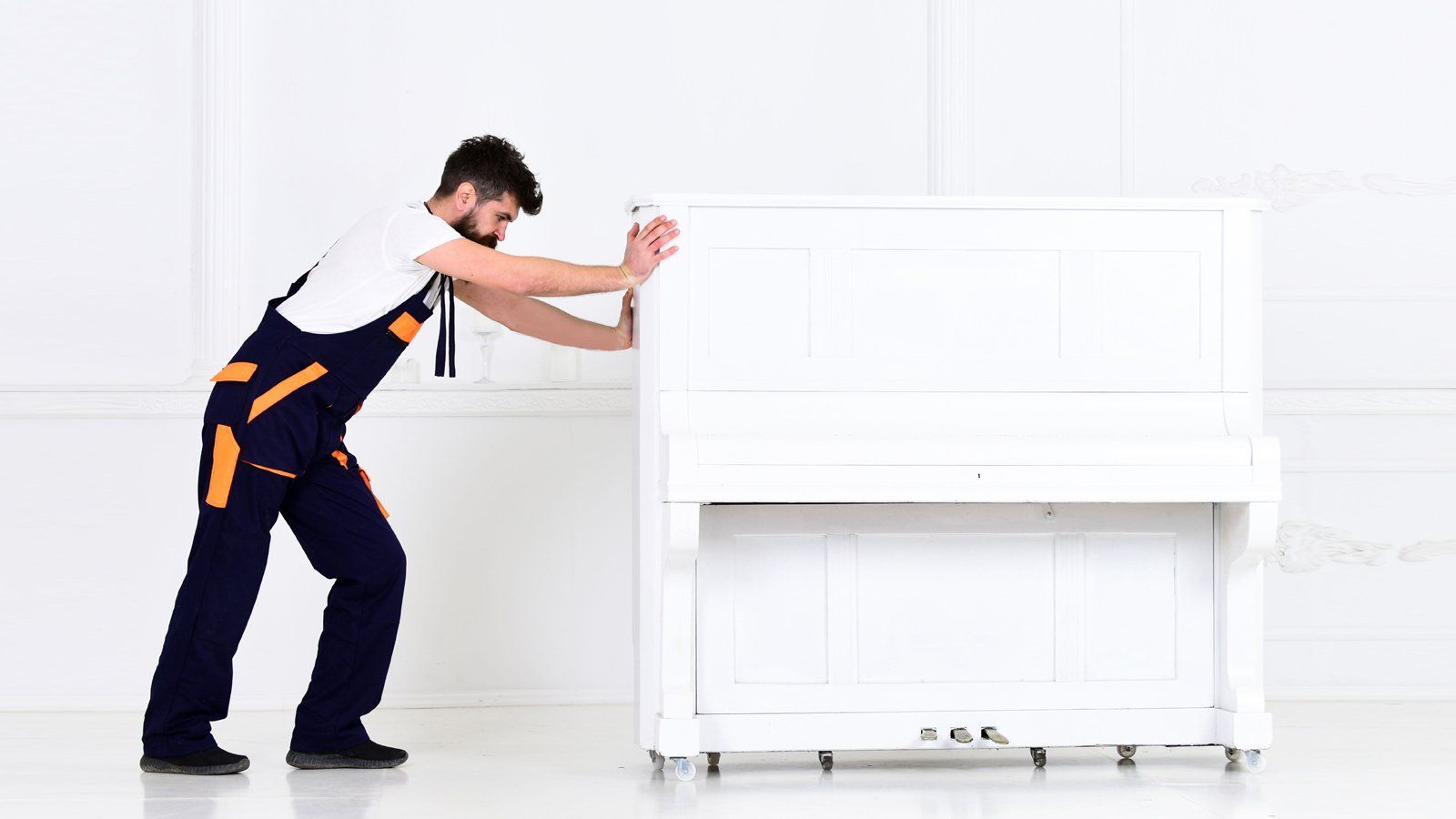 Man in blue overalls pushes a white piano against a white wall.