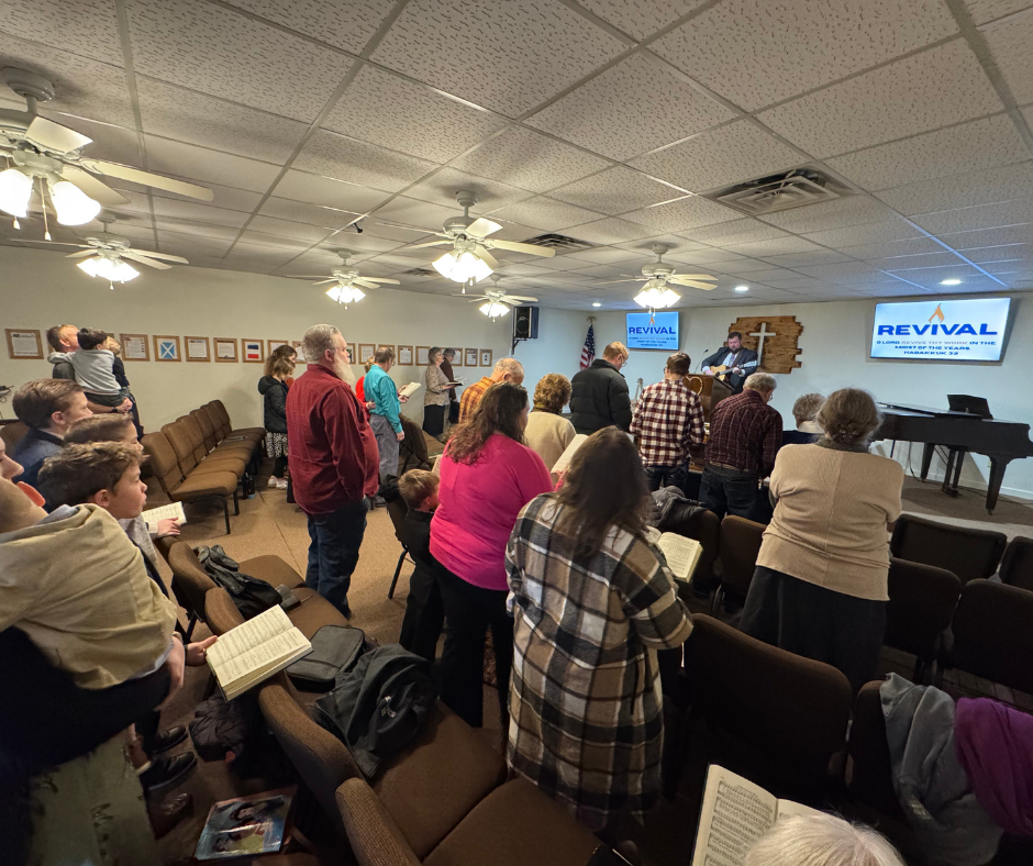 Interior view of a church sanctuary with rows of pews, a stage with a cross, and multiple ceiling fans.