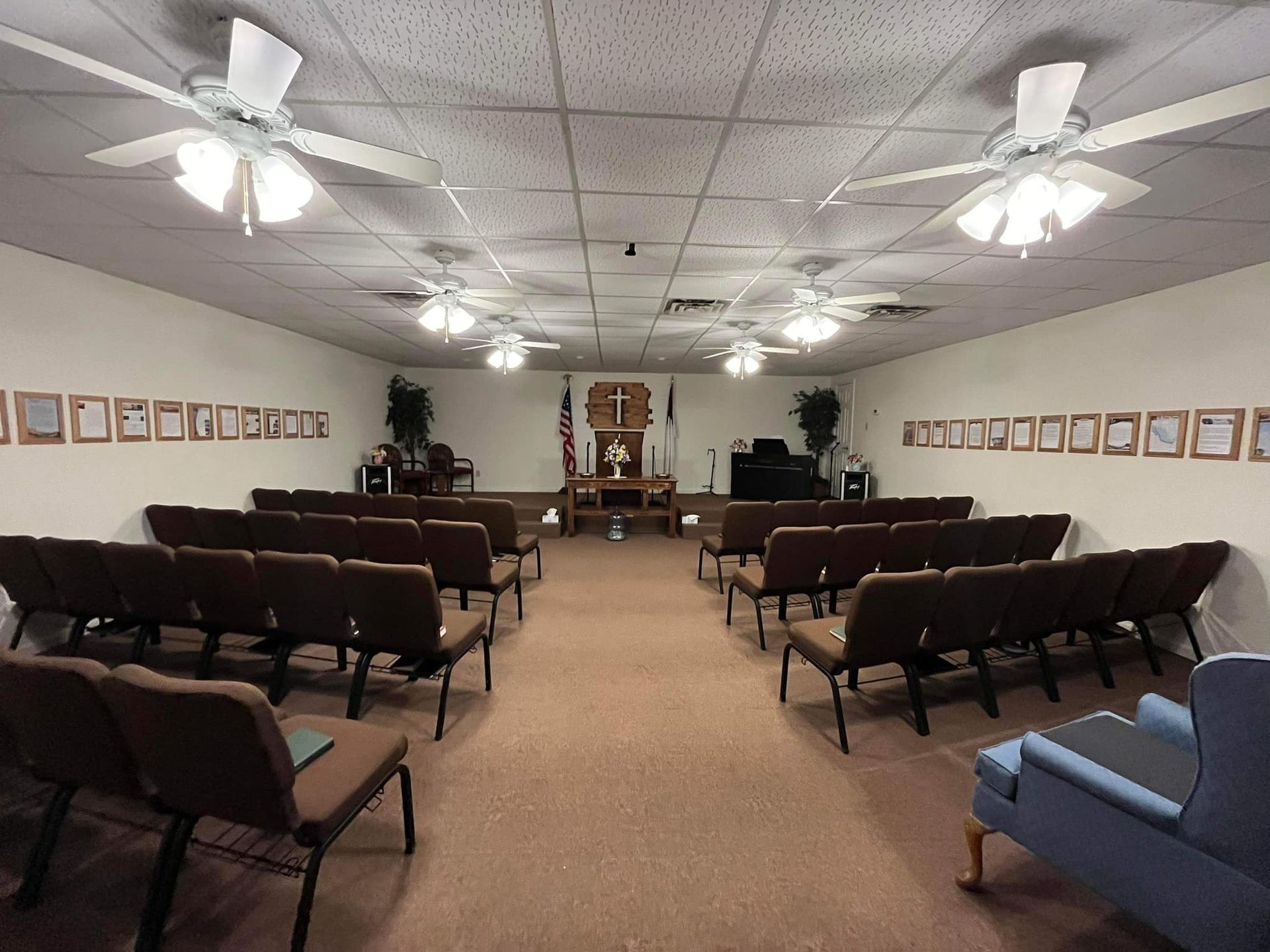 Interior view of a church sanctuary with rows of pews, a stage with a cross, and multiple ceiling fans.