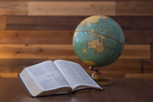 Opened Bible next to a globe on a wooden table, against a wooden wall background.