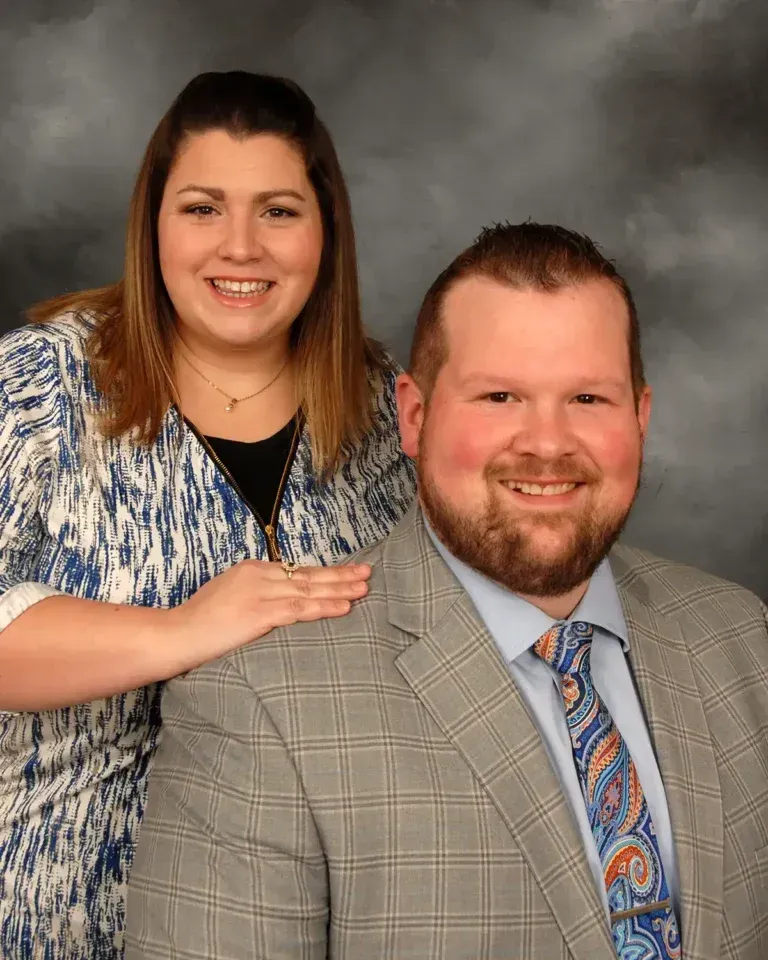 Woman with hand on the shoulder of a man in a plaid jacket. Both smiling, against a blurred gray backdrop.