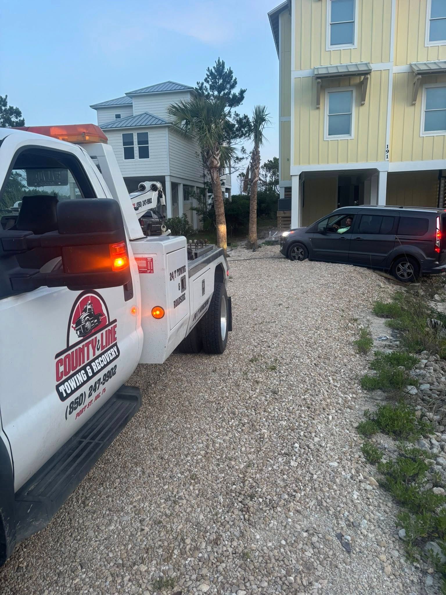 Tow truck in gravel driveway beside a yellow house, car being towed.