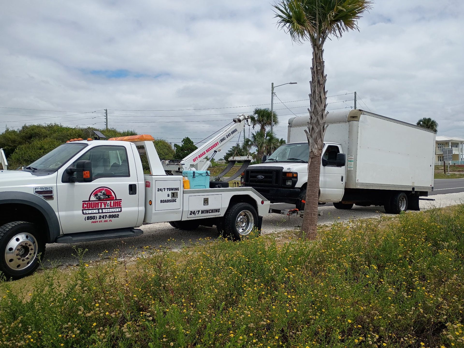 Tow truck towing a white box truck. Outdoors, sunny day.