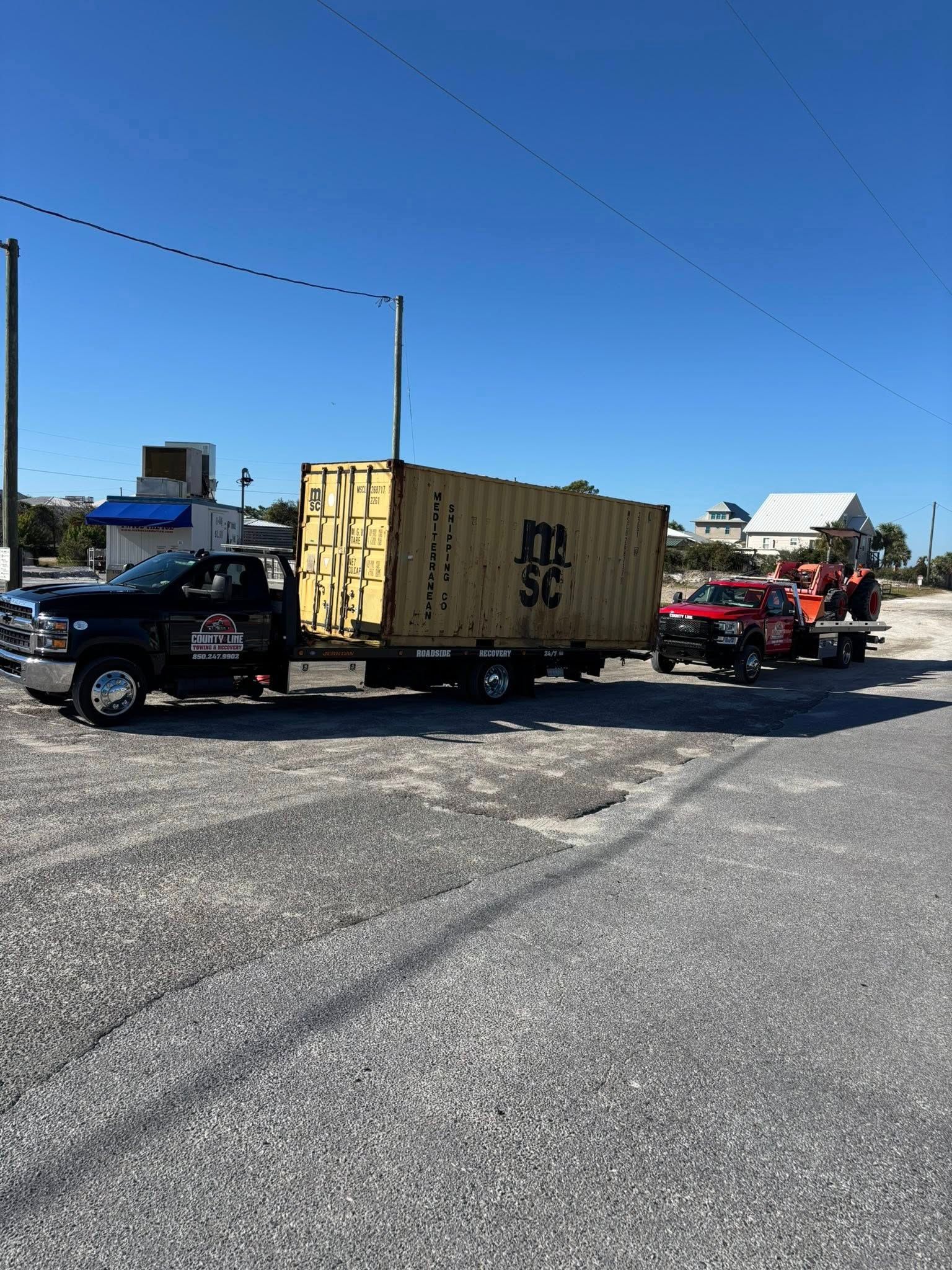 Two trucks towing a large shipping container on a gravel lot under a clear blue sky.