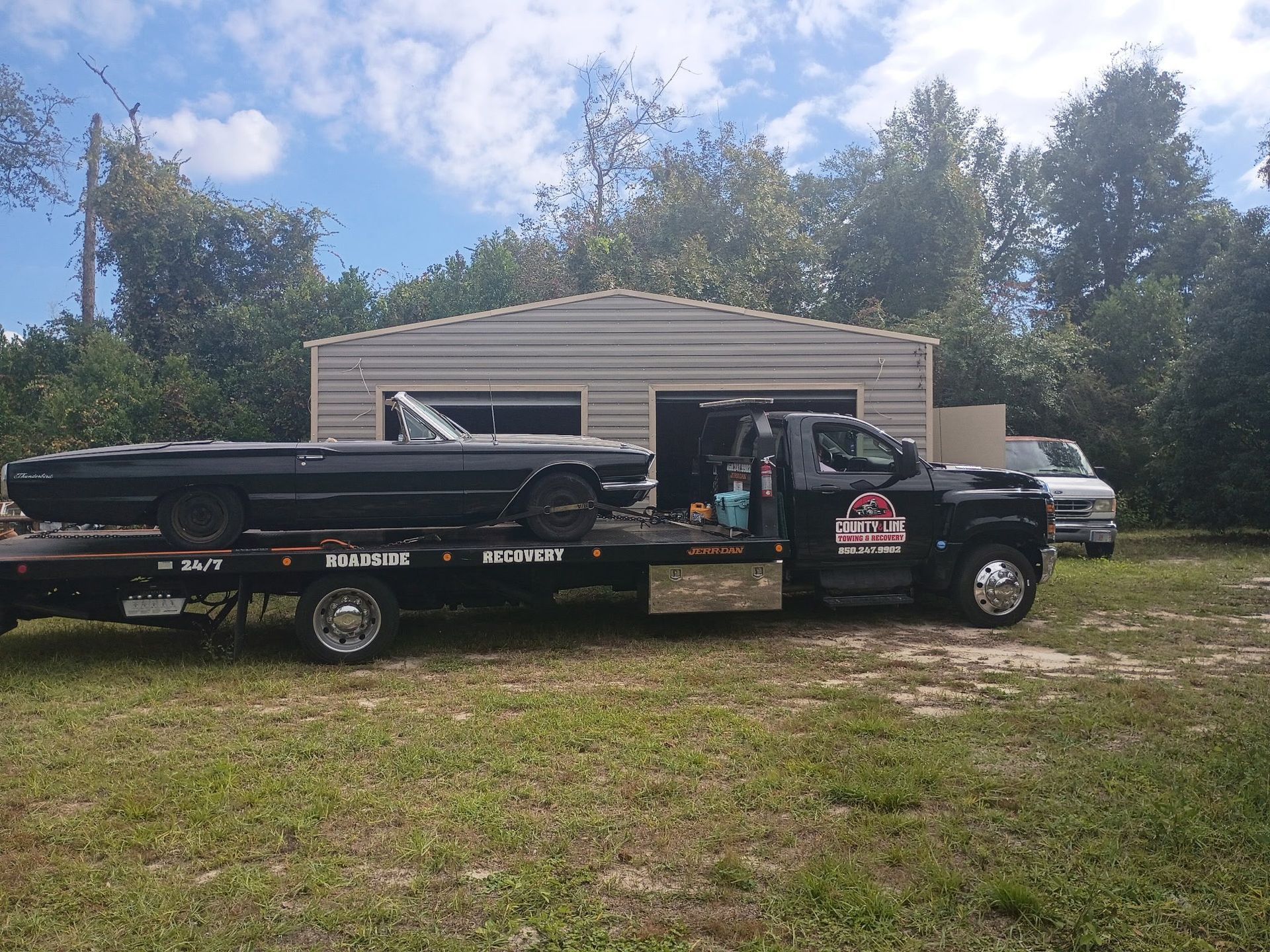 Black convertible car on a flatbed tow truck. Truck is parked in front of a garage on a grassy area.