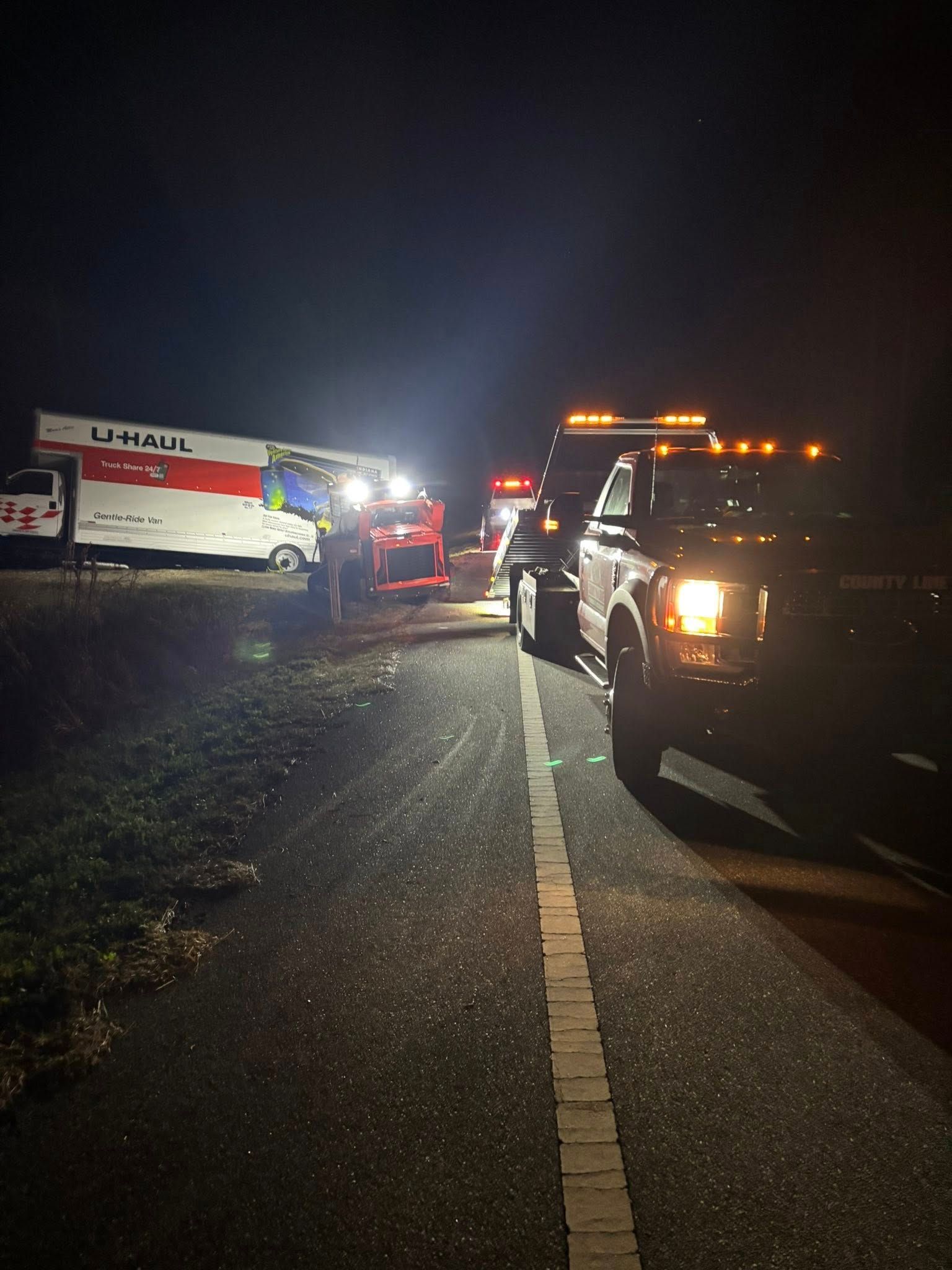 Tow truck and other vehicles working on a U-Haul truck at night on the side of a highway.