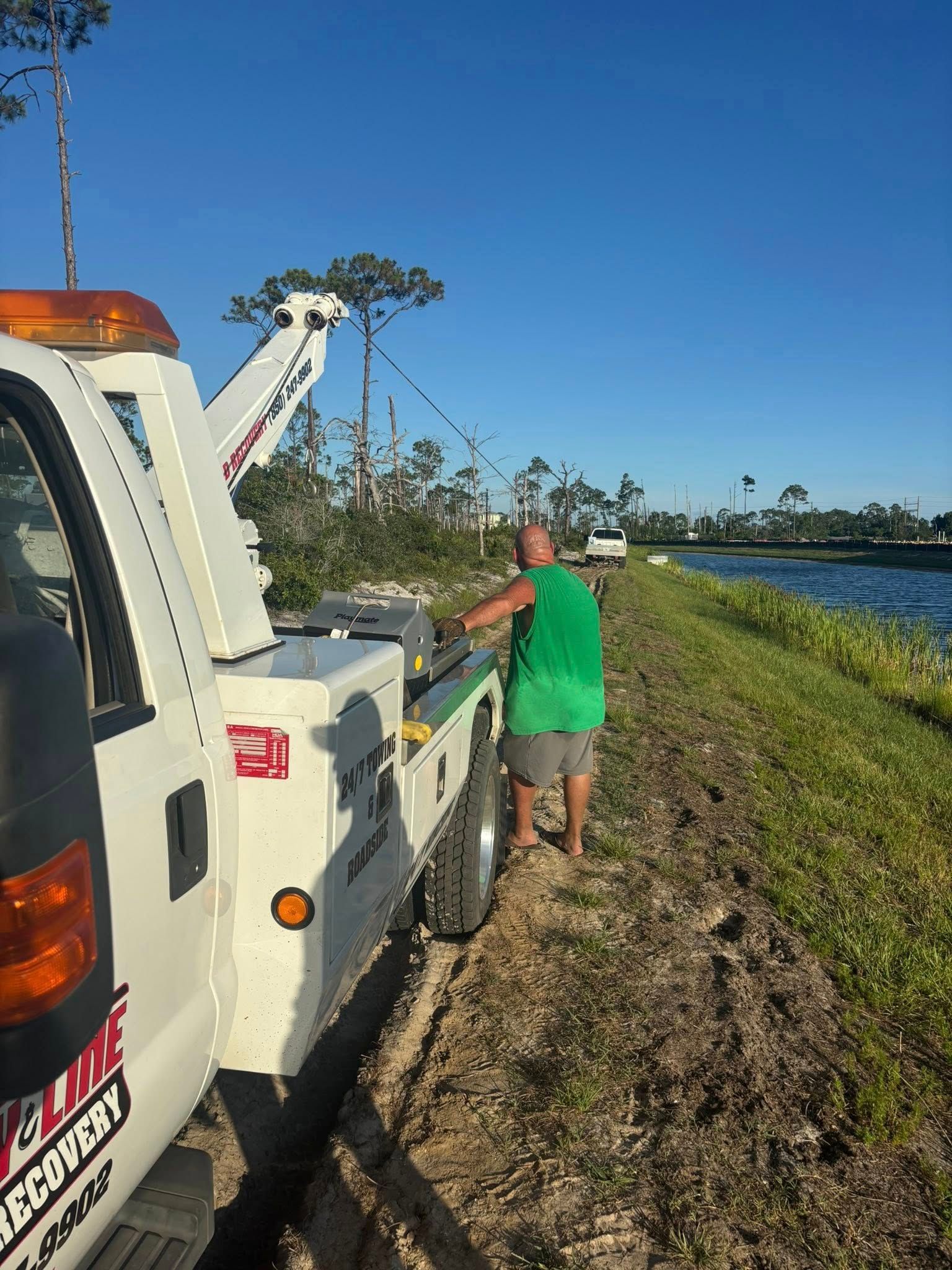 Tow truck pulling a vehicle from a grassy ditch near water. Man in green shirt stands by the truck.