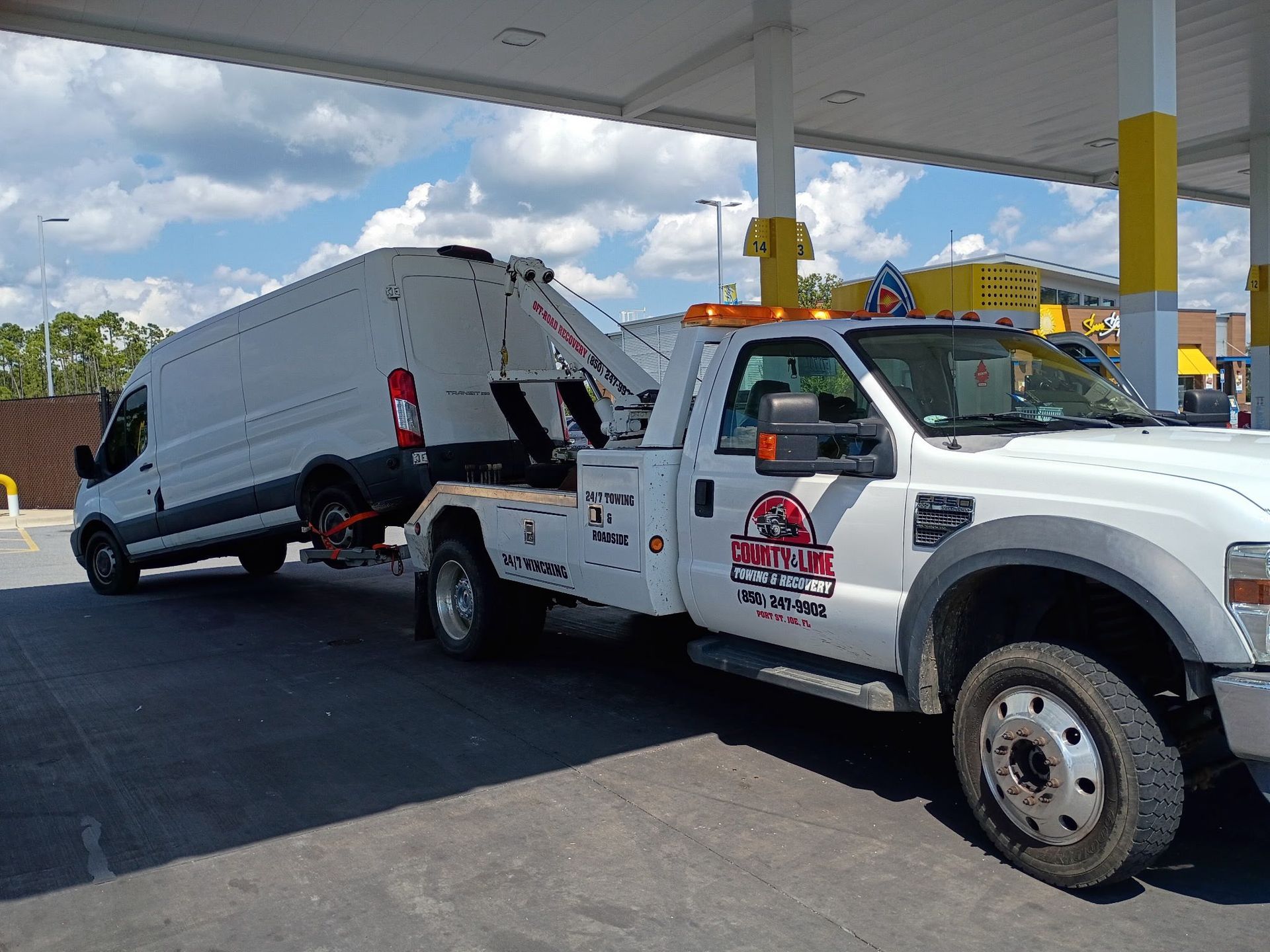 White tow truck towing a white cargo van at a gas station under a blue sky.